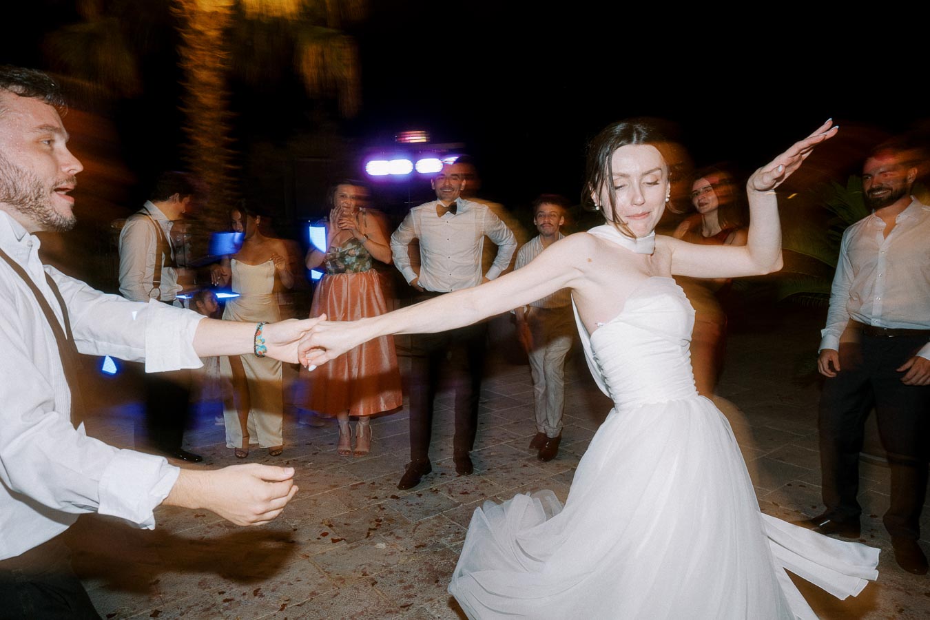 Wedding dance celebration with bride in white gown joyfully dancing with guests at nighttime, surrounded by friends in formal attire under colorful lights.