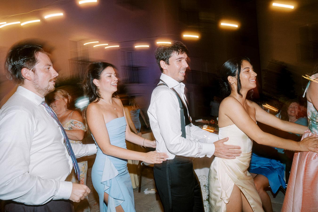 A group of people joyfully dancing in a conga line at a lively outdoor evening event, dressed in formal attire with blurred lights creating a festive atmosphere in the background.