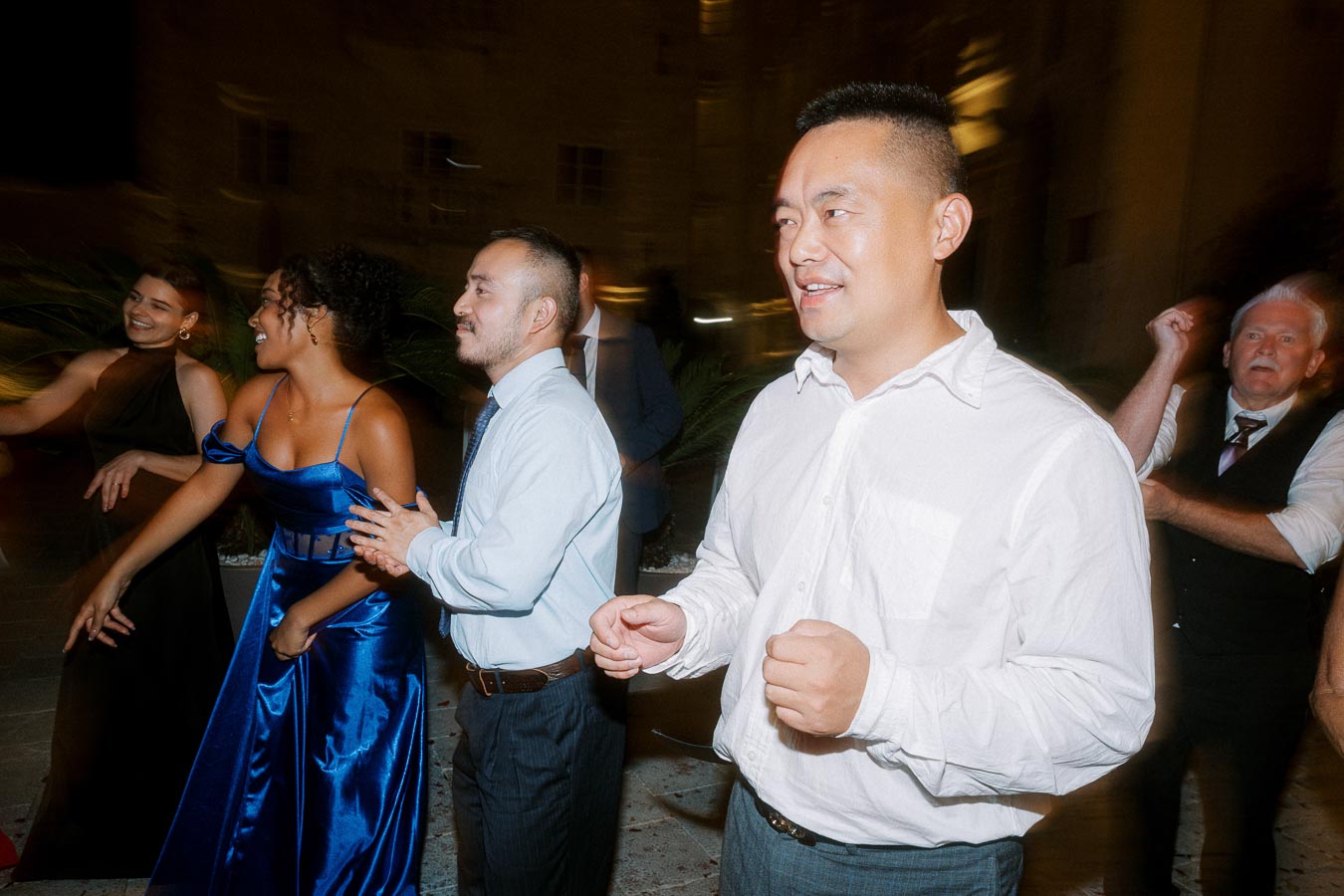 A group of people joyfully dancing at a night event, dressed in formal attire including elegant dresses and shirts, surrounded by a vibrant party atmosphere.
