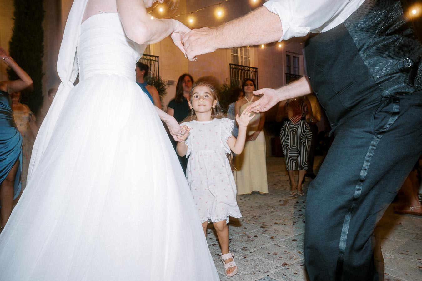 A young girl dances with a bride and groom at an outdoor evening wedding reception, surrounded by guests, with string lights in the background.