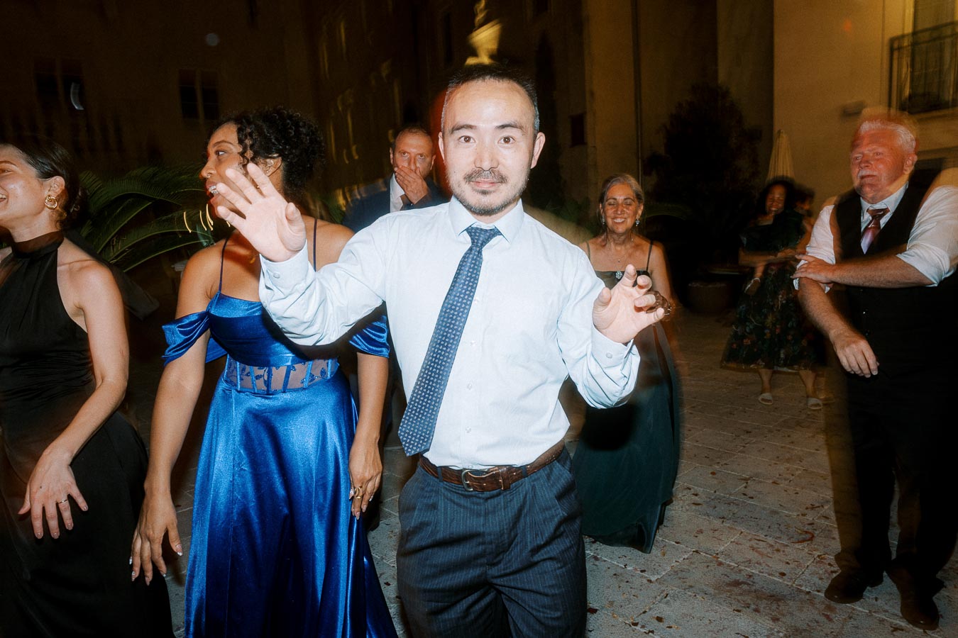 A group of people in formal attire dancing joyfully at an outdoor evening event, with a man in a white shirt and tie in the foreground waving hands.