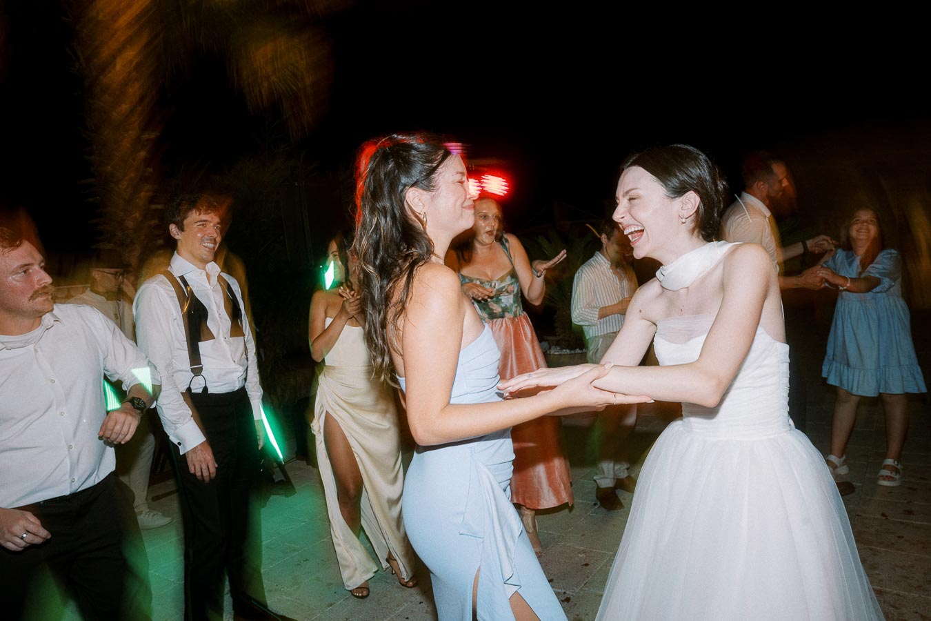 A group of people joyfully dancing at a wedding celebration under evening lighting, with one woman in a white dress sharing a moment of laughter with another woman in a light blue dress, surrounded by well-dressed guests enjoying the event.