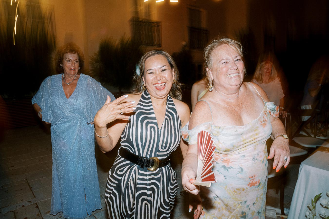Three women joyfully smiling and dancing in elegant dresses at a lively evening party, one holding a red fan, under soft ambient lighting.