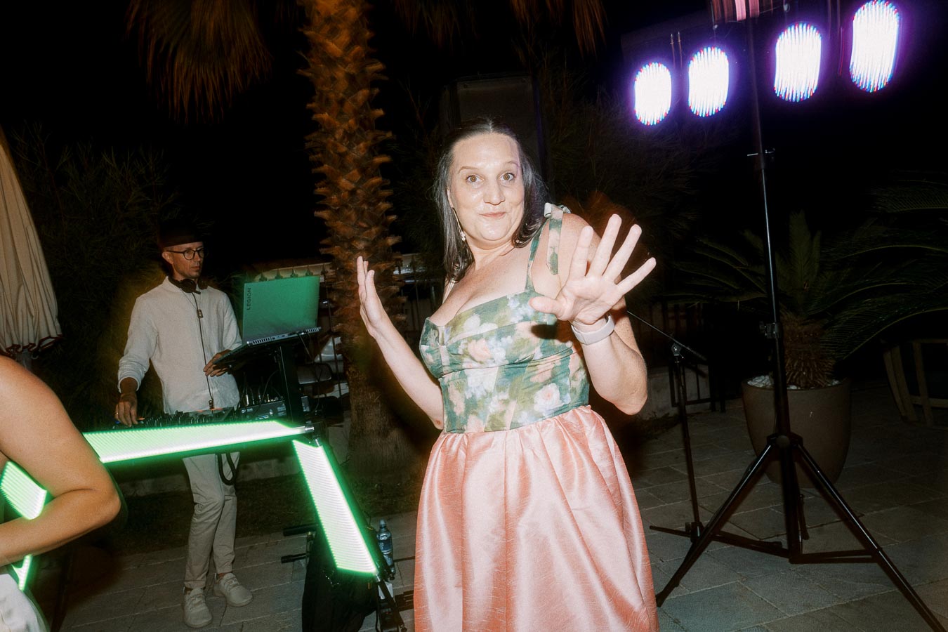 A woman in a floral dress dancing at an outdoor night event with a DJ in the background, colorful lights illuminating the scene.