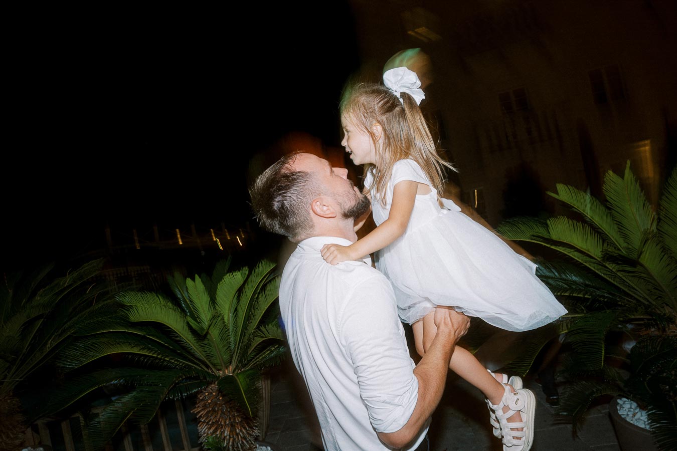 Father lifting daughter in white dress at night, surrounded by palm plants.