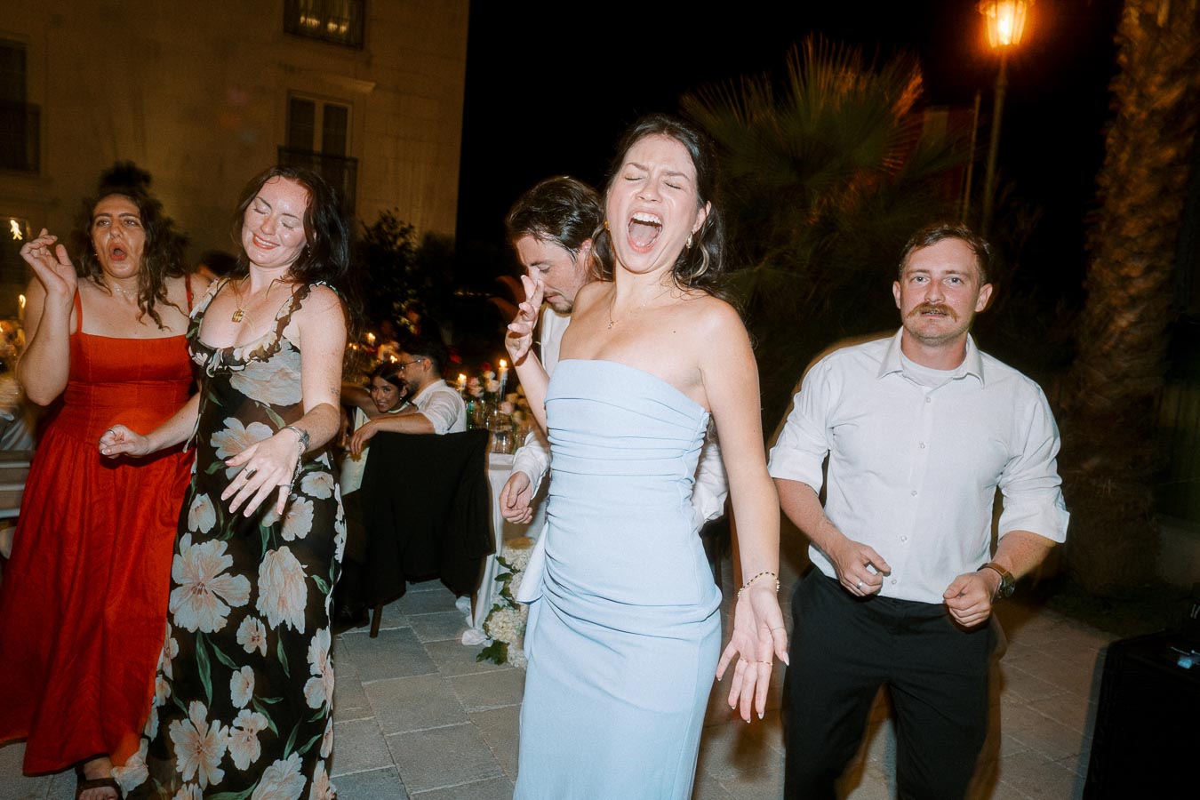 A group of people joyfully dancing at an outdoor evening event, with one woman in a blue dress laughing exuberantly, surrounded by others in vibrant attire.