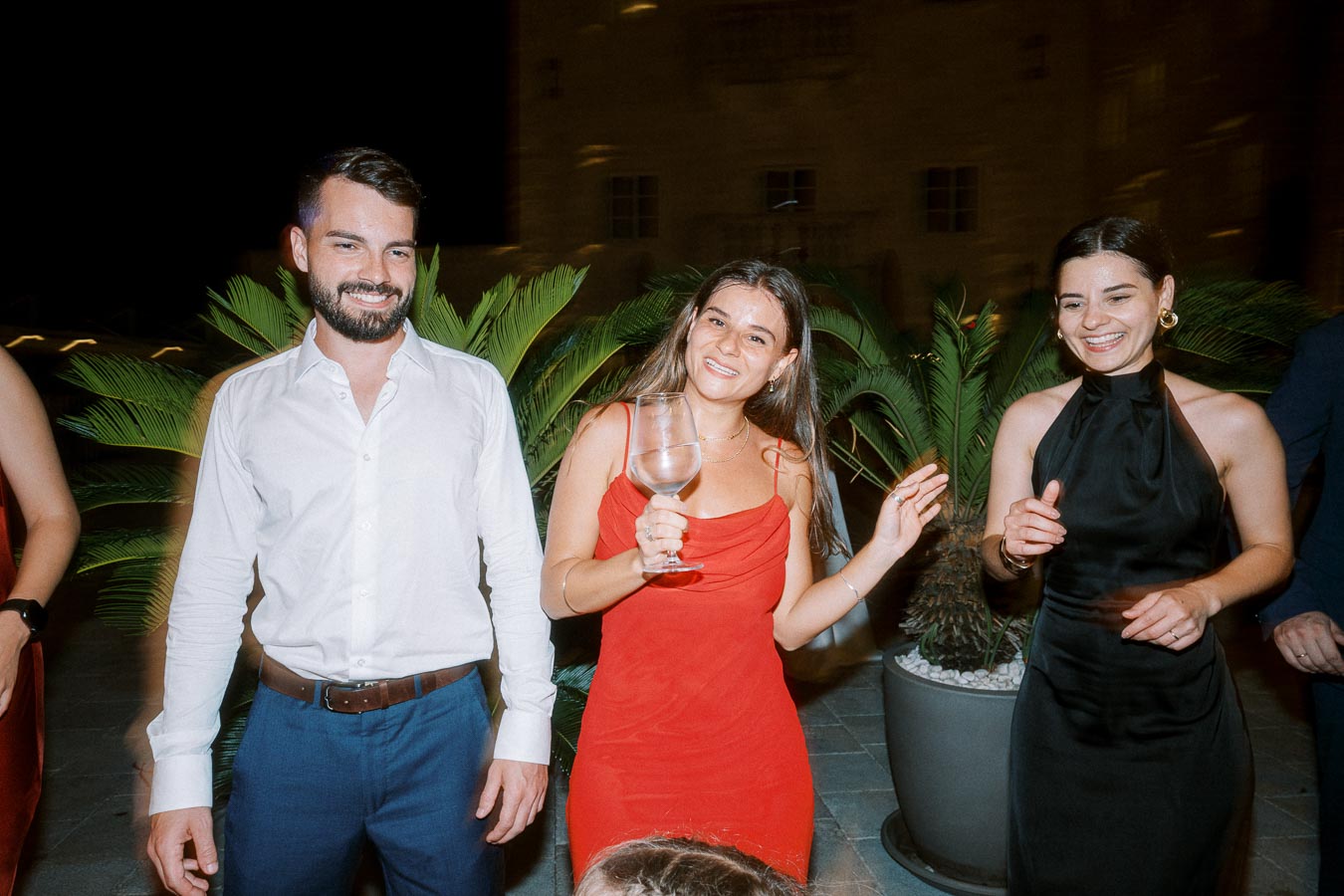 Group of people enjoying a nighttime party, with women in elegant dresses and a man in a white shirt, standing in front of palm plants, smiling and holding a drink.