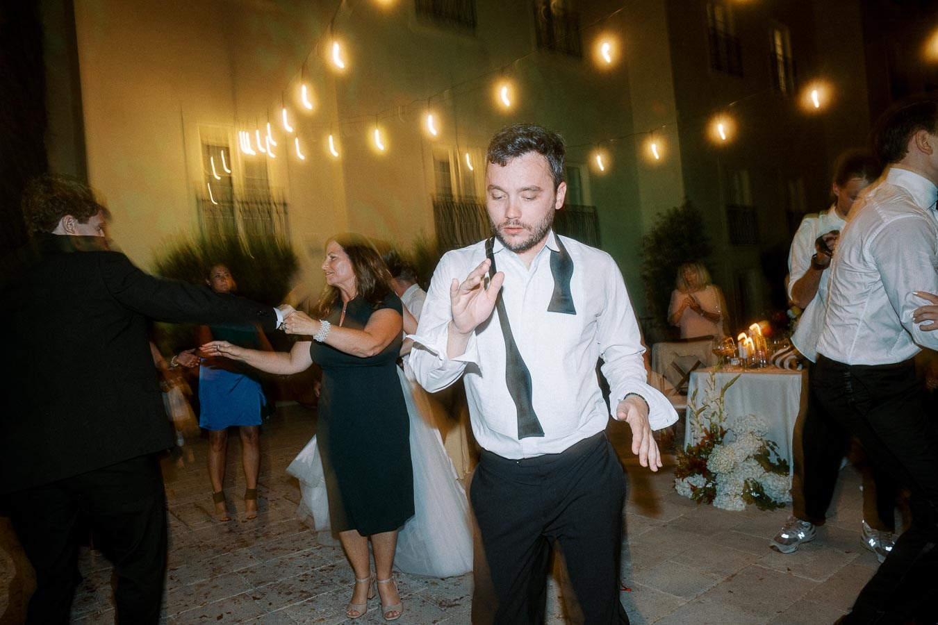 A man in a white shirt with an untied black tie dances energetically at an outdoor evening event, surrounded by other guests in formal attire and string lights.