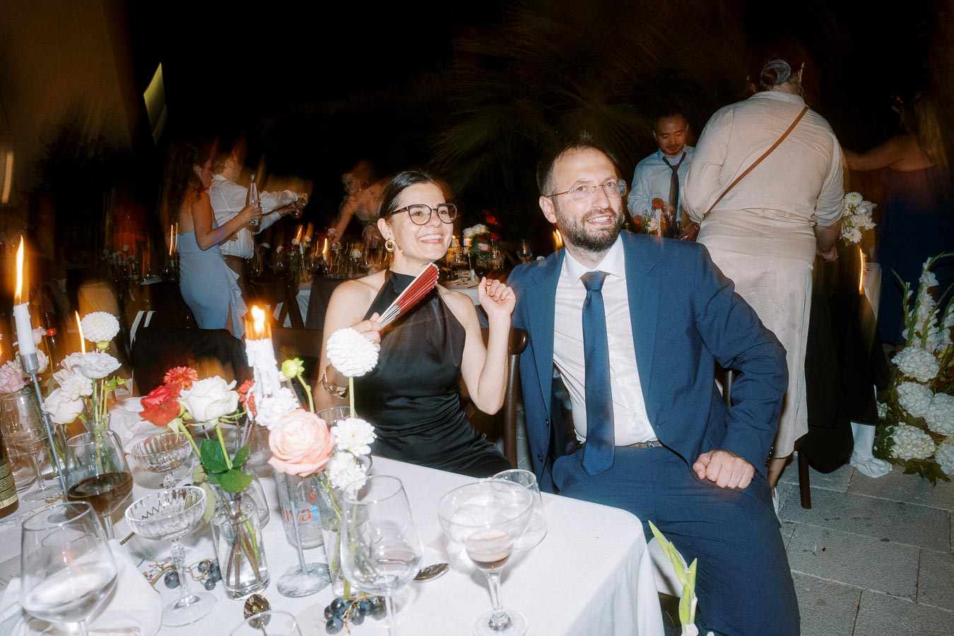 A well-dressed couple enjoying an elegant outdoor evening event, sitting at a beautifully decorated table with floral arrangements and candlelight, surrounded by other guests socializing in the background.