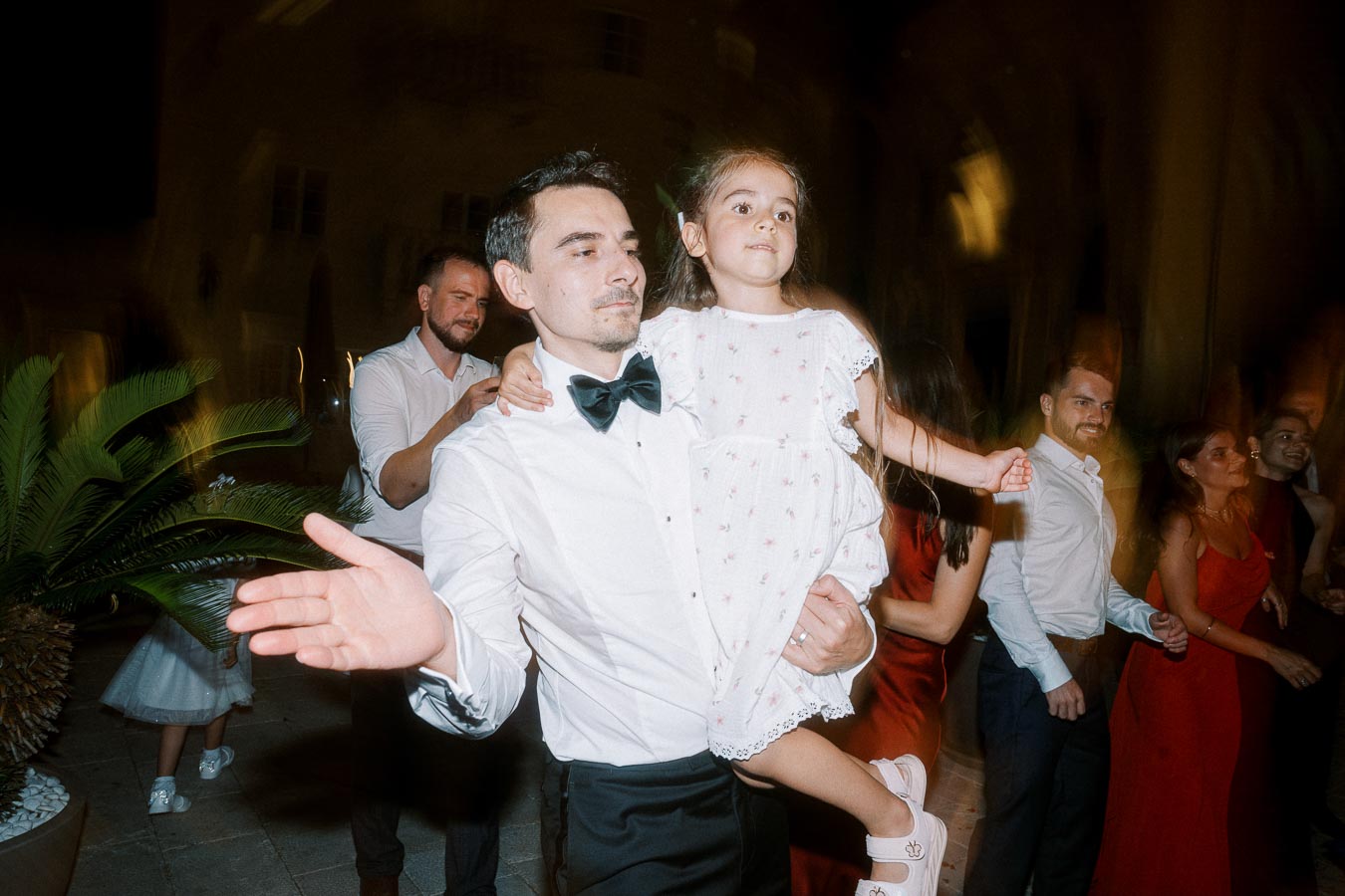 A man in a white shirt and black bow tie holds a young girl in a white dress while dancing at an indoor wedding reception, surrounded by other guests in formal attire.