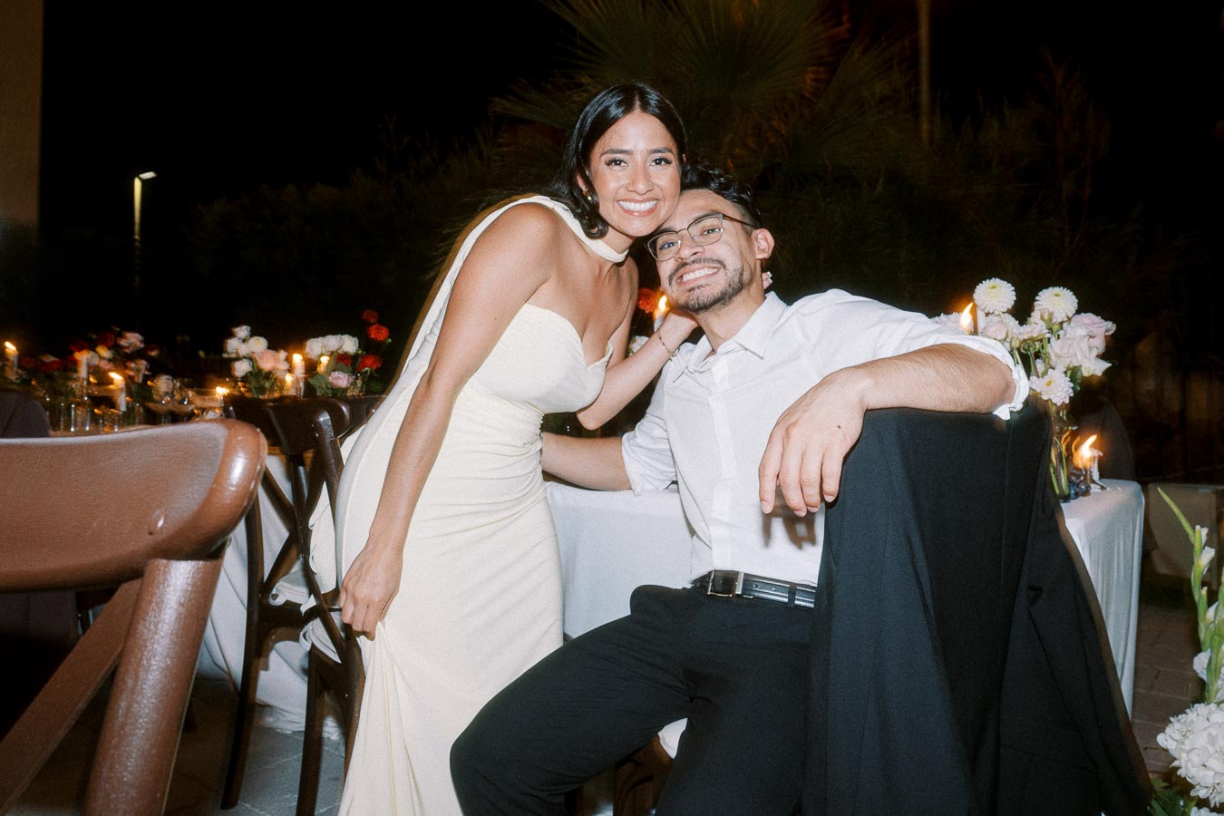 A smiling couple poses at a candlelit outdoor evening event, with a table decorated with flowers and candles in the background.