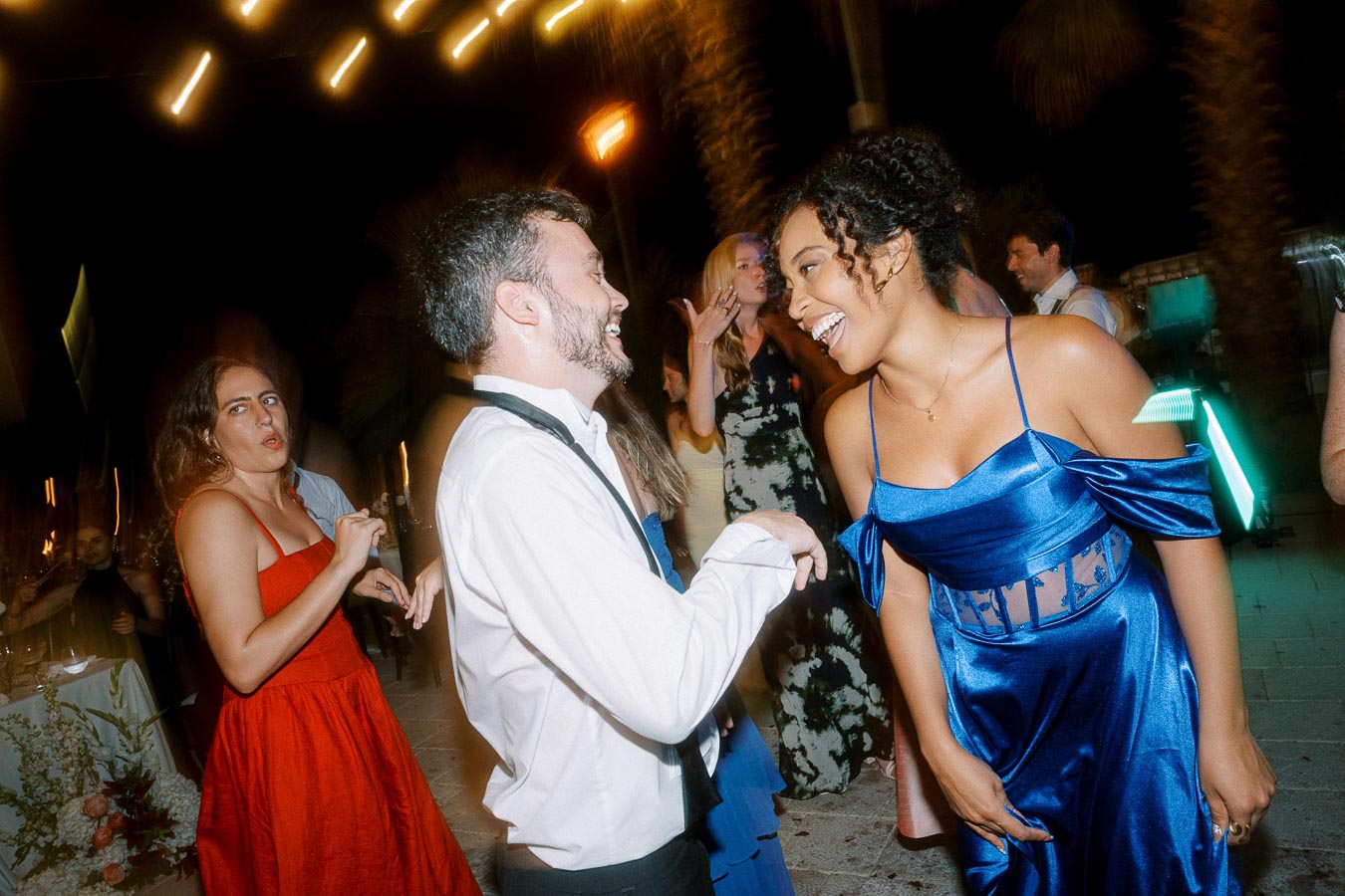 People joyfully dancing at a nighttime celebration, with one person in a red dress and another in a blue dress, surrounded by festive lighting and other guests in formal attire.