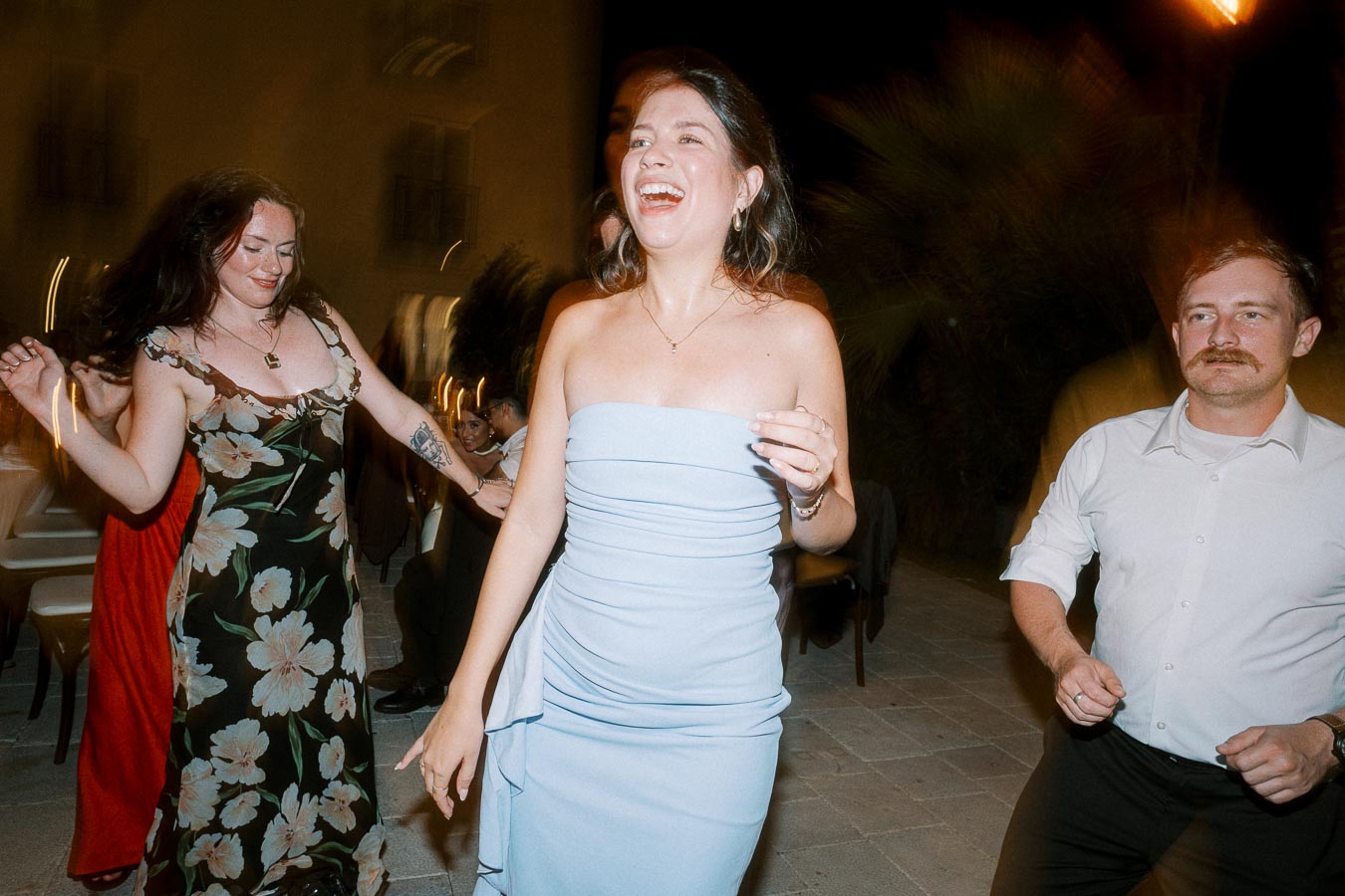 Three people are joyfully dancing at an evening party. The woman in the center wears a light blue strapless dress and is laughing, while a woman in a floral dress and a man in a white shirt join in the festivities. The dim setting suggests a lively night event.