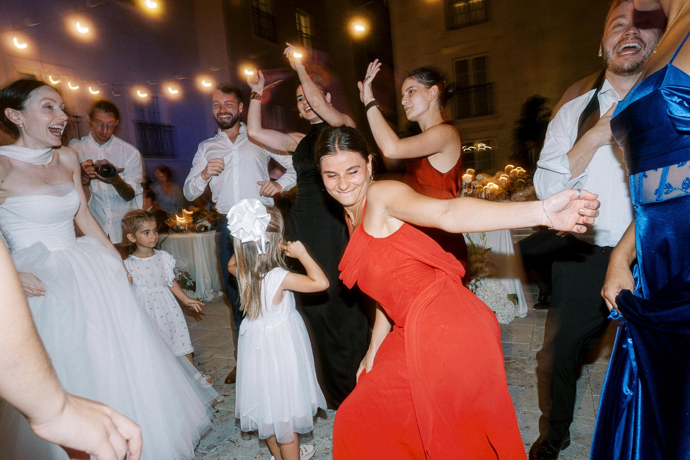 Wedding reception with a joyful bride in a white dress and guests dancing exuberantly in colorful outfits, set against a backdrop of string lights and nighttime ambiance.