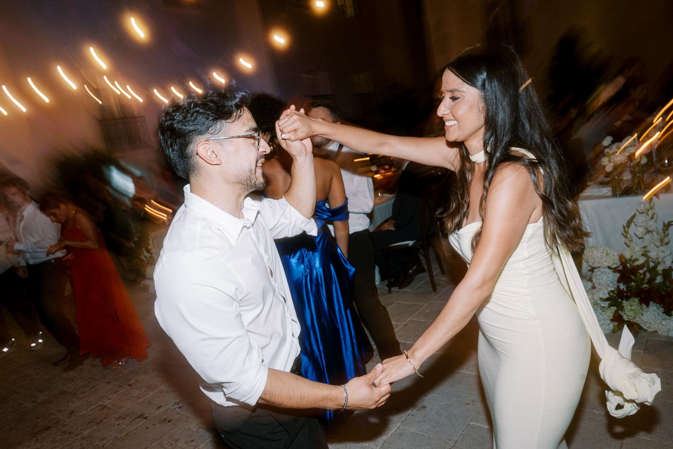 A man and woman joyfully dancing together at a festive evening event, surrounded by blurred lights and guests in elegant attire.