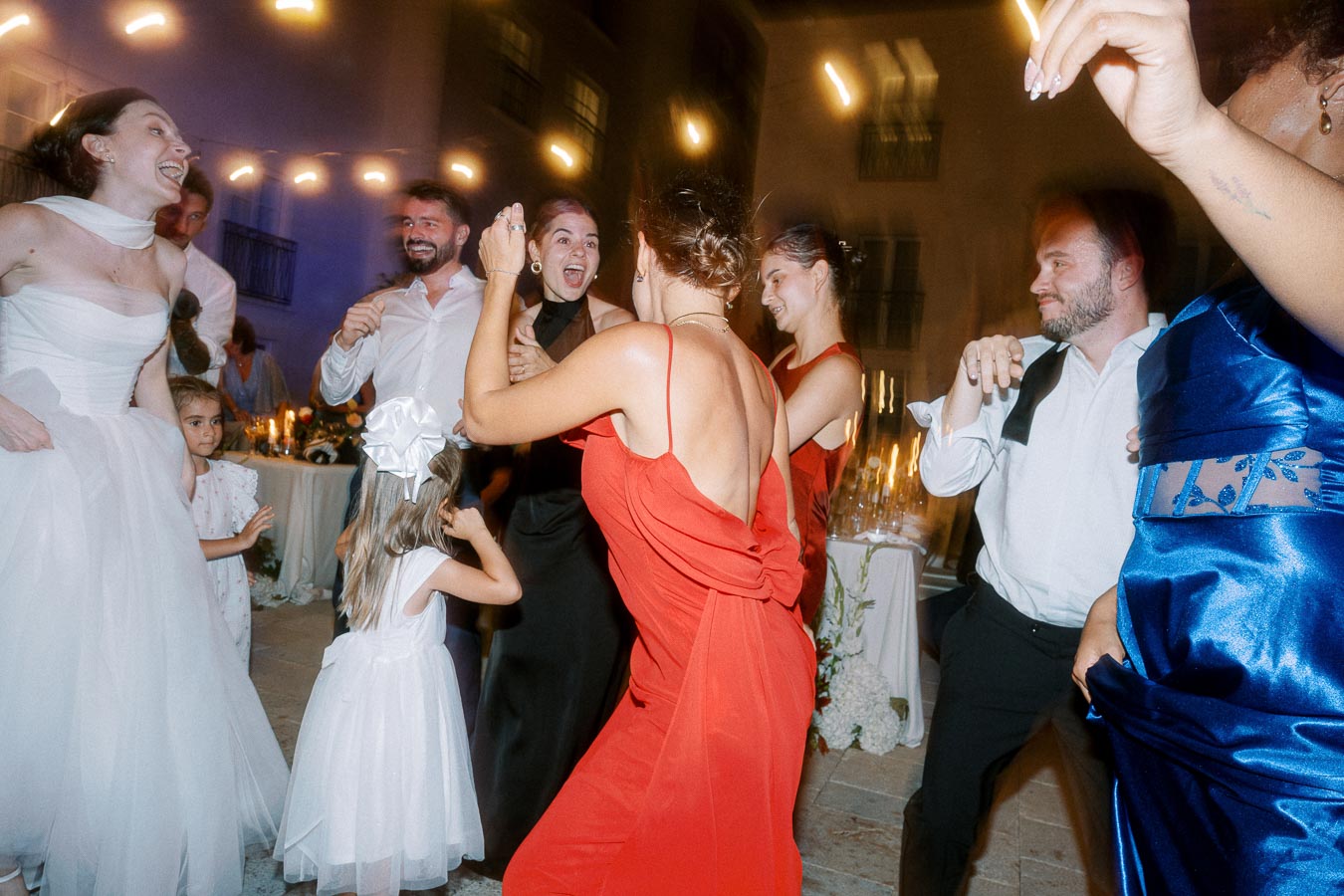 The image shows a joyful group of people dancing at a wedding reception. The bride in a white dress and other guests in elegant attire, including red and blue dresses, are celebrating together under ambient lights. A young girl in a white dress with a bow is also visible, contributing to the festive atmosphere.