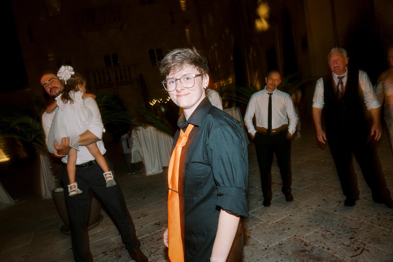 A person wearing a black shirt and orange tie at an evening event, surrounded by a group of people, including one holding a child, in a festive setting.