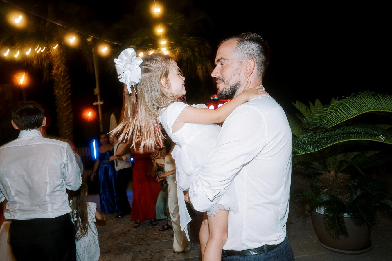 A father in a white shirt joyfully holds his young daughter, who is wearing a white dress with a large hair bow, at an outdoor evening event decorated with string lights and palm plants.