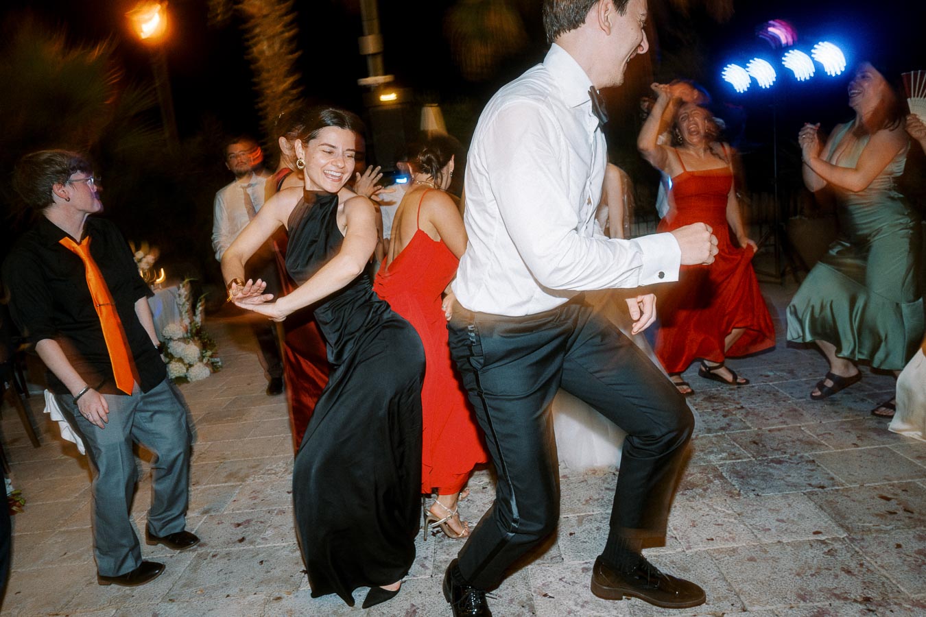 Group of people joyfully dancing at a lively outdoor party at night, wearing elegant formal attire, under colorful lights.