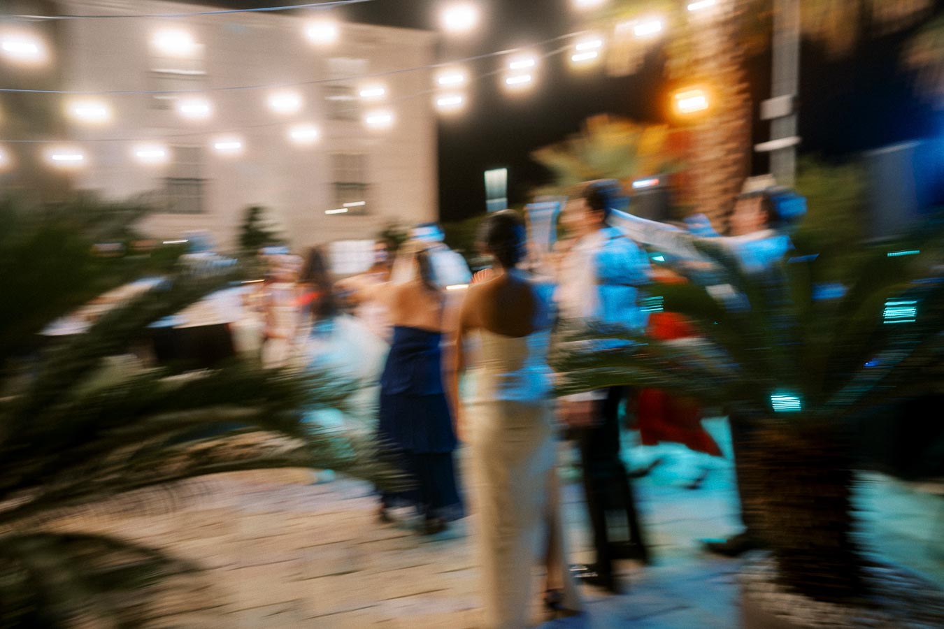 Blurred image of an outdoor evening event with string lights above and people mingling around a palm plant, creating a festive atmosphere.