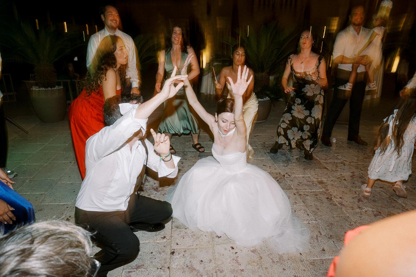 A joyful bride in a white gown dances energetically with guests at an outdoor evening wedding reception.