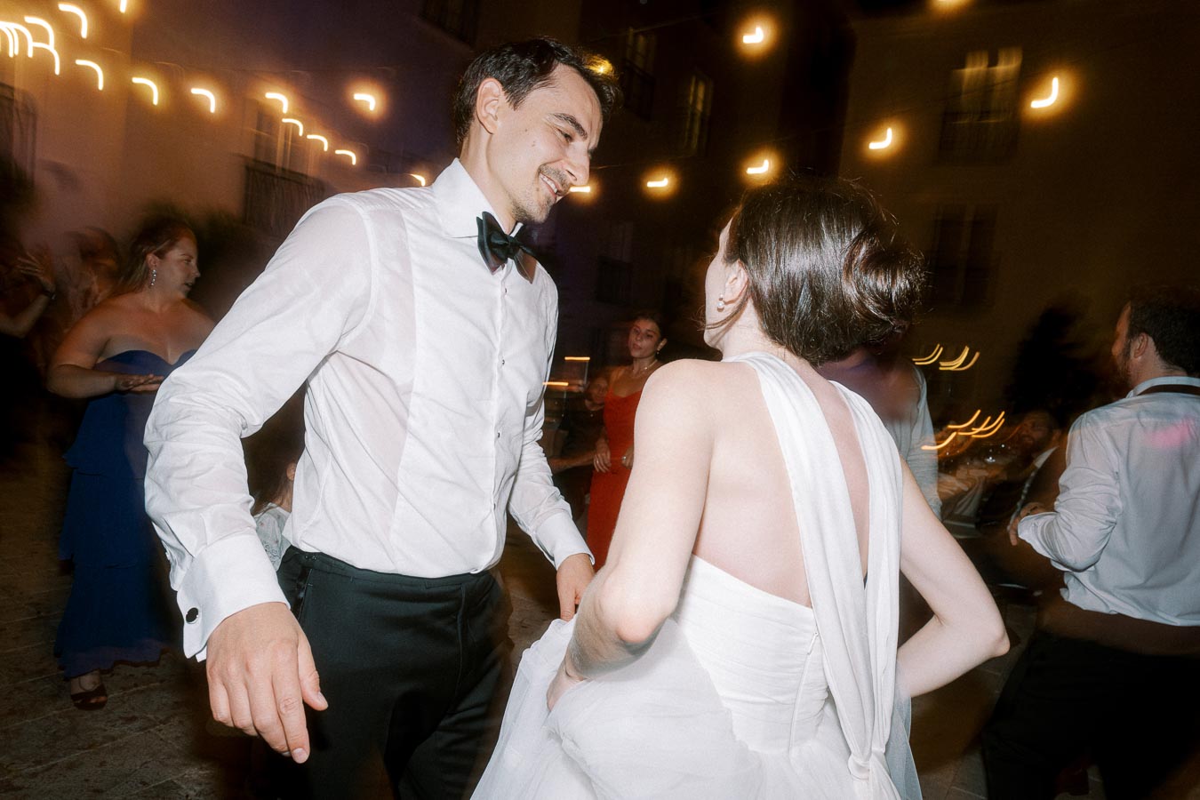 A couple dancing at a wedding reception, surrounded by guests in formal attire and illuminated by string lights creating a festive atmosphere.