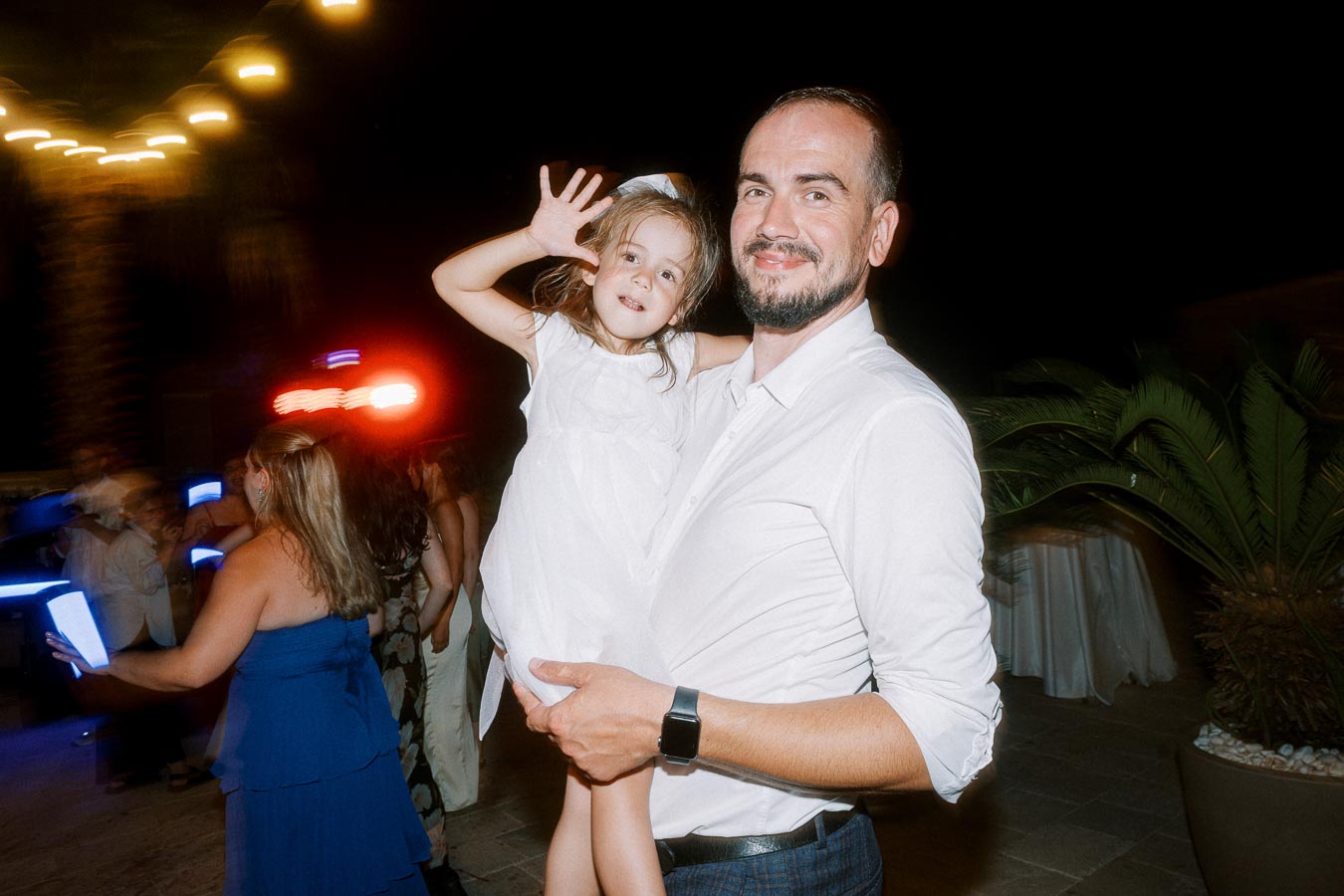 A man in a white shirt holds a young girl wearing a white dress, both smiling at an outdoor evening party with blurred lights and attendees in the background.