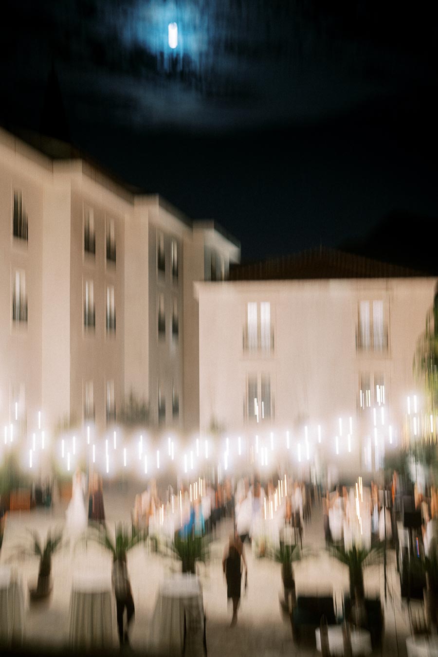 Blurry evening wedding reception outdoors with string lights, white tables, and guests amidst a backdrop of elegant buildings under a moonlit sky.