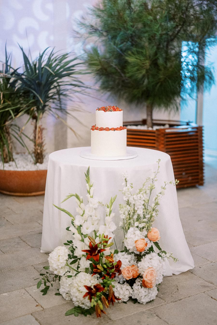 Two-tier white wedding cake with orange floral decoration, set on a round, white-draped table, adorned with white hydrangeas, orange roses, and greenery, surrounded by lush potted plants in an elegant indoor setting.