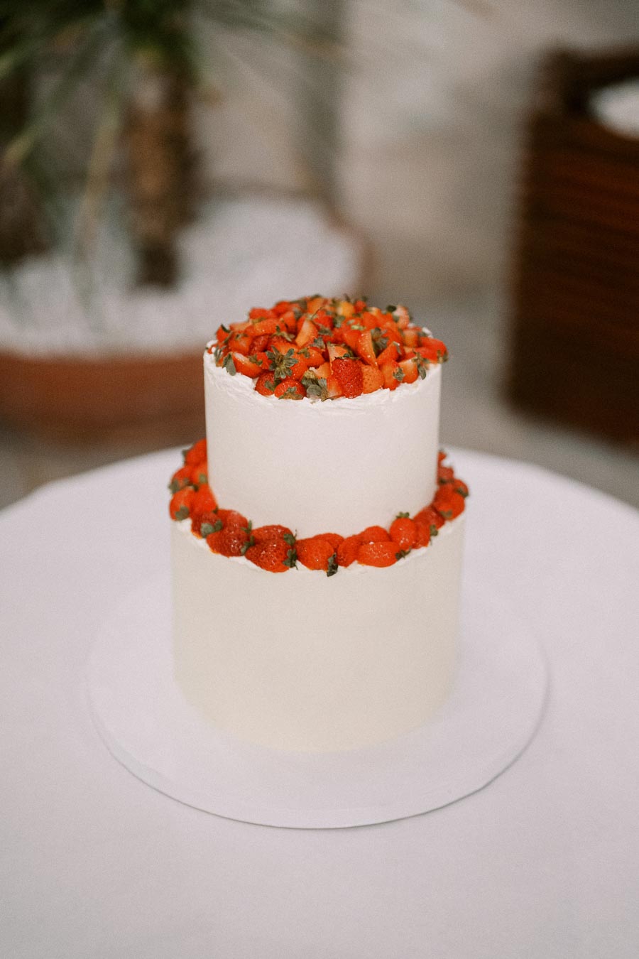 Two-tiered white cake topped with fresh strawberries and surrounded by a strawberry border, displayed on a white tablecloth.