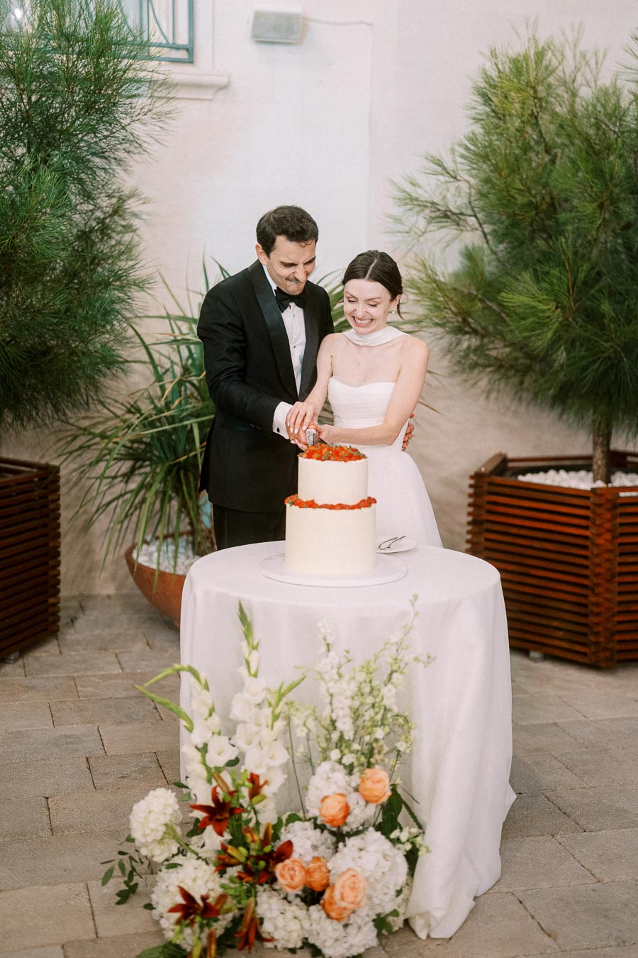 A couple in formal attire cuts a white wedding cake adorned with flowers on a table, surrounded by greenery and floral arrangements.