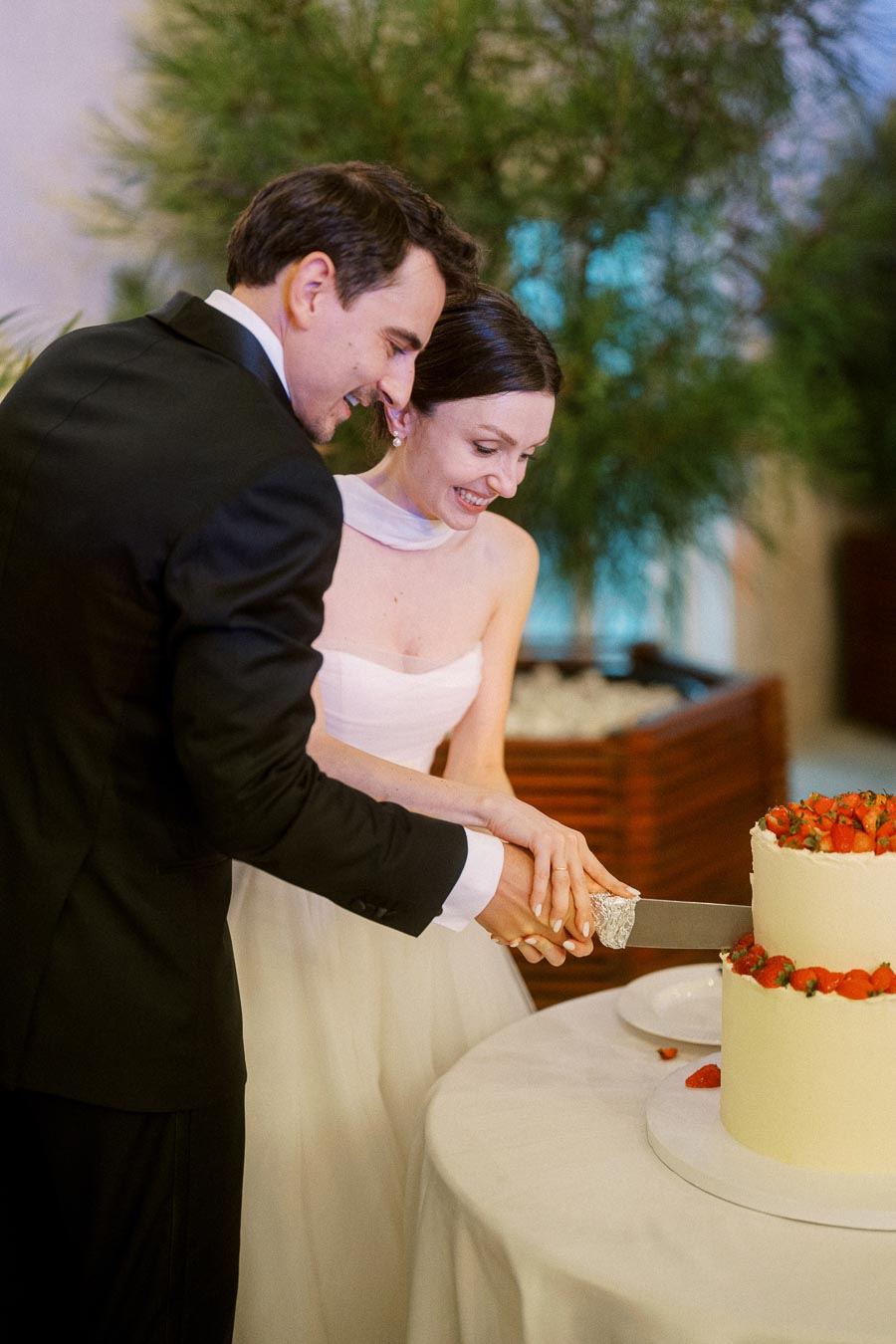 A bride and groom smiling as they cut their wedding cake together, adorned with fresh strawberries, at a reception.