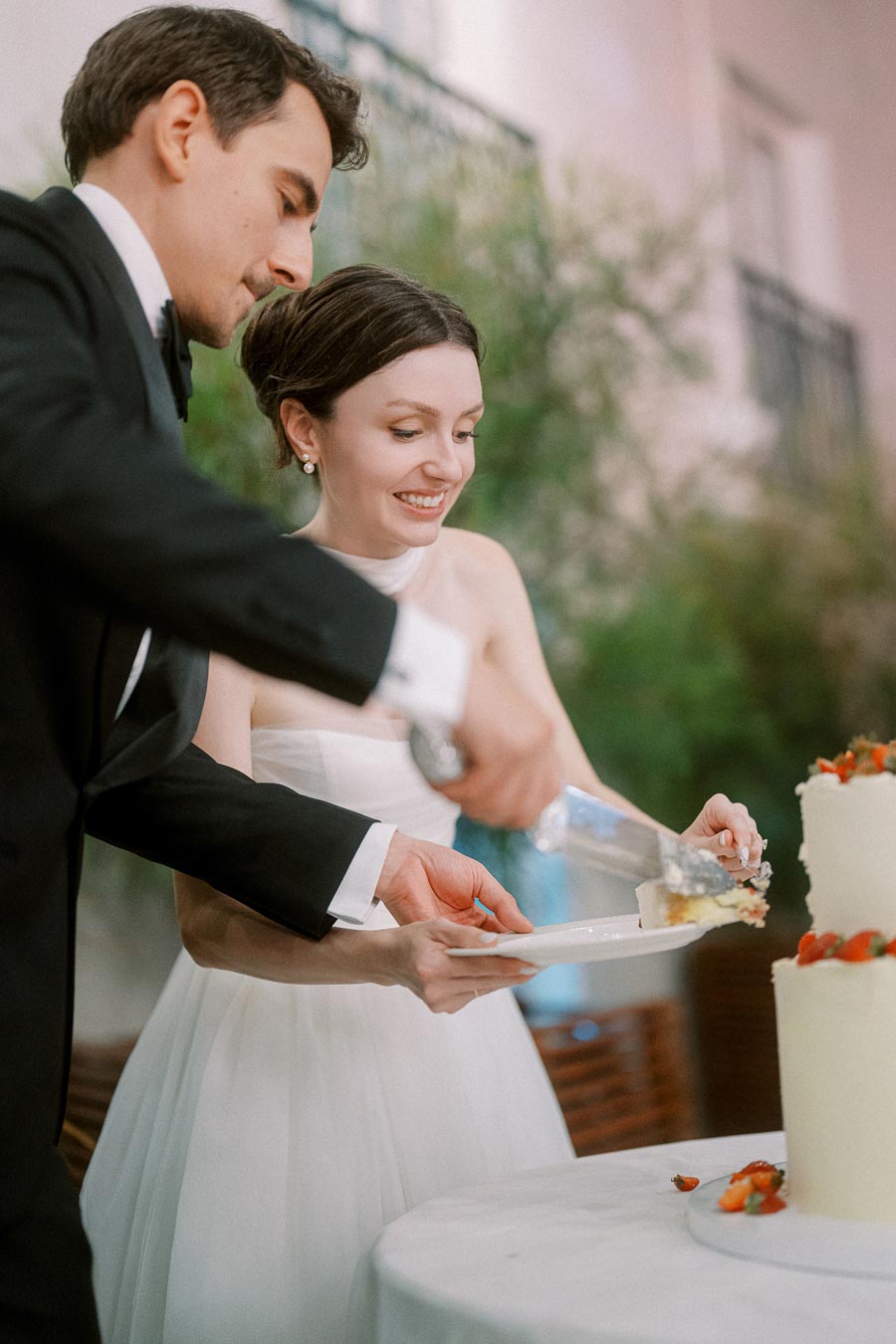 A bride and groom in formal attire cutting a three-tiered white wedding cake adorned with fresh strawberries, smiling as they serve a slice onto a plate during their wedding celebration.