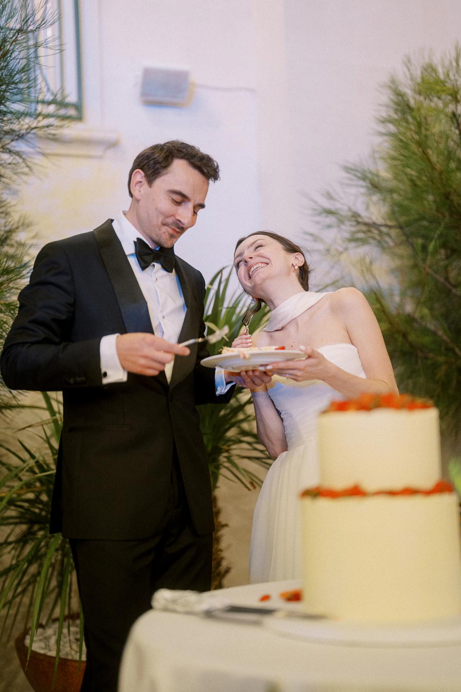A joyful couple in wedding attire shares a lighthearted moment while cutting their wedding cake, surrounded by lush greenery.