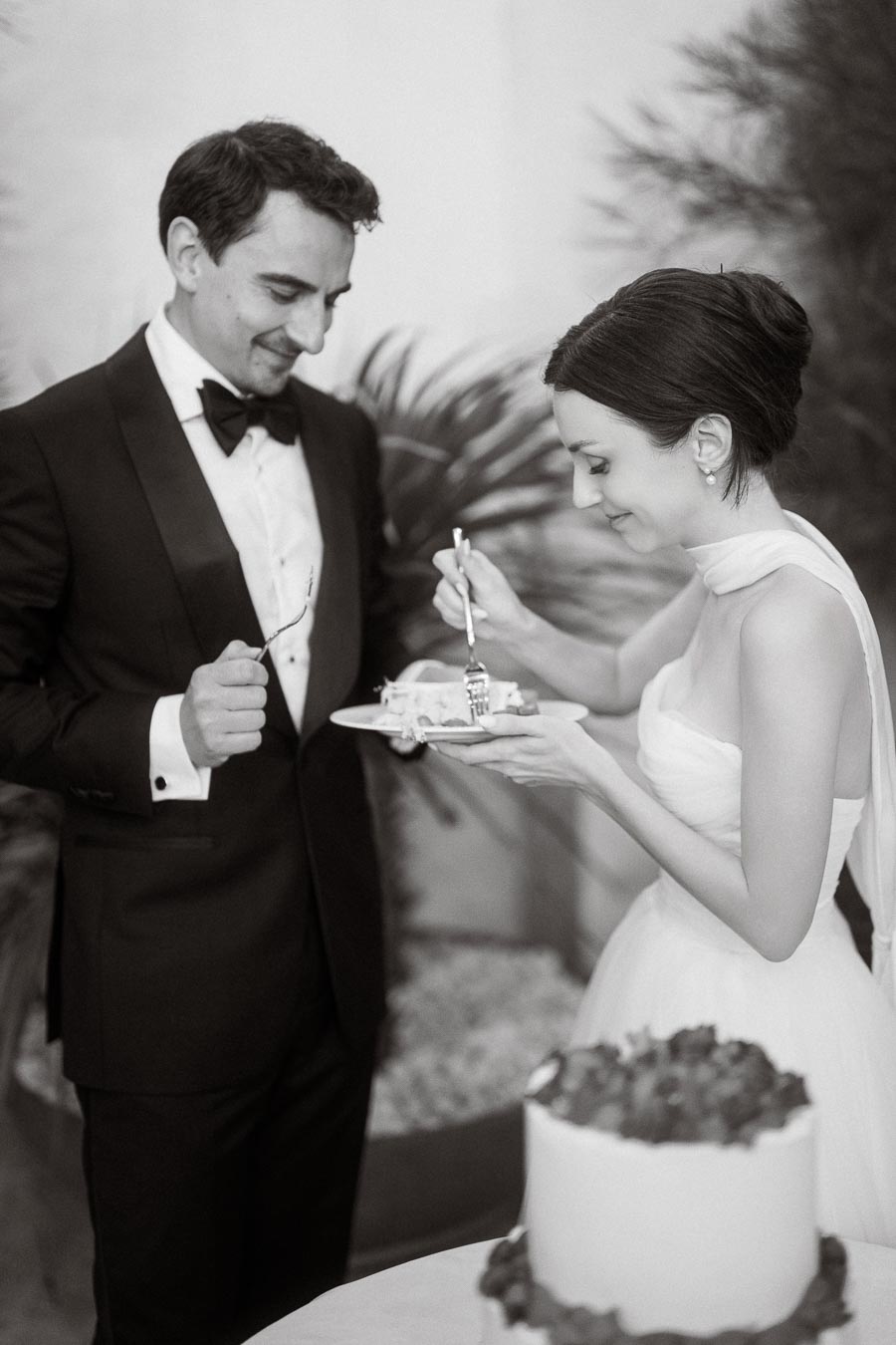 A bride and groom enjoy a slice of wedding cake together in a black and white photo, highlighting their joyful moment and elegant attire.