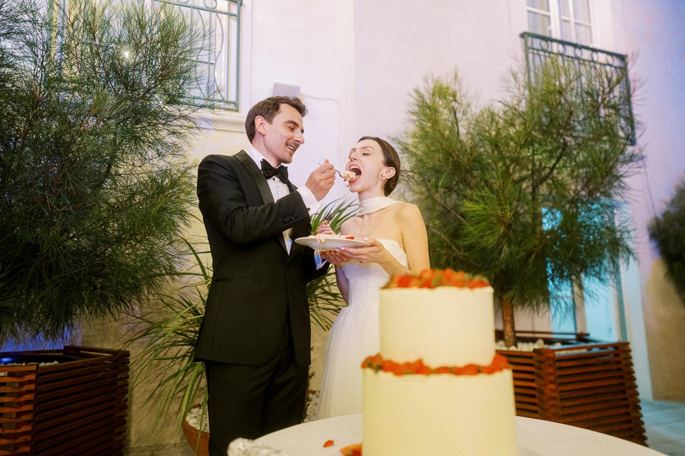 A bride and groom in formal attire smiling as they feed each other wedding cake outdoors, with a decorated cake in the foreground and greenery in the background.
