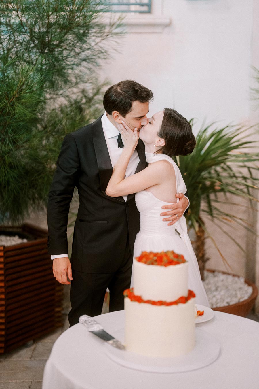 A bride and groom share a romantic kiss next to a white wedding cake adorned with red flowers, surrounded by lush greenery at an elegant outdoor venue.