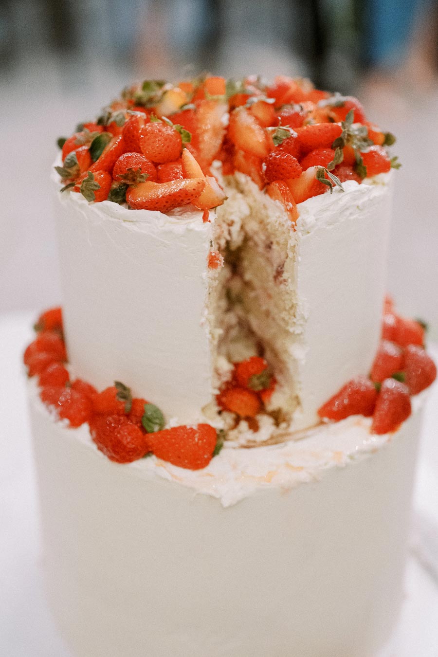 Two-tiered white cake with a slice cut out, topped with fresh strawberries, showcasing a creamy interior and scattered berries around the base.