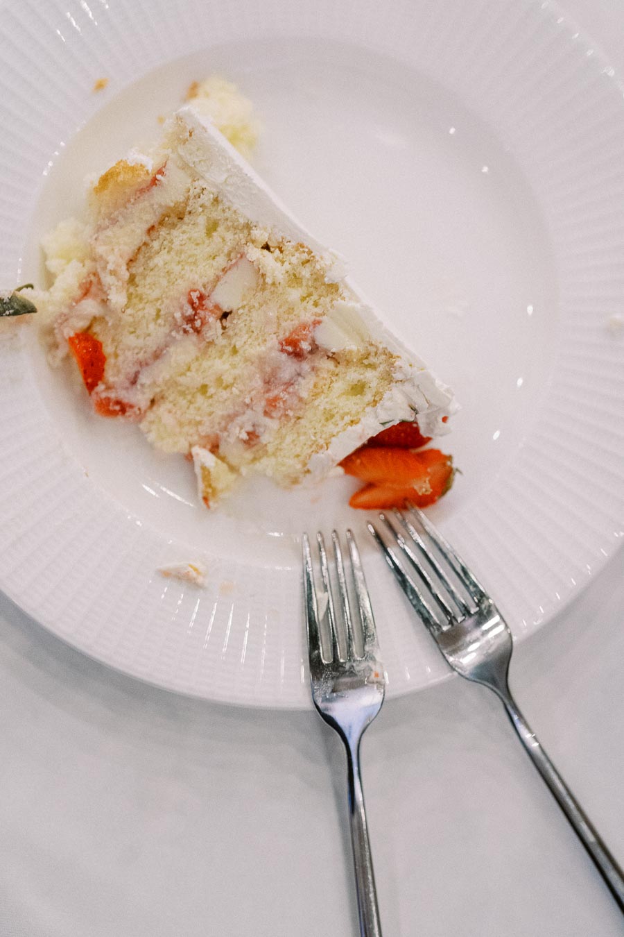 A slice of strawberry layer cake with cream frosting on a white plate, accompanied by two silver forks.