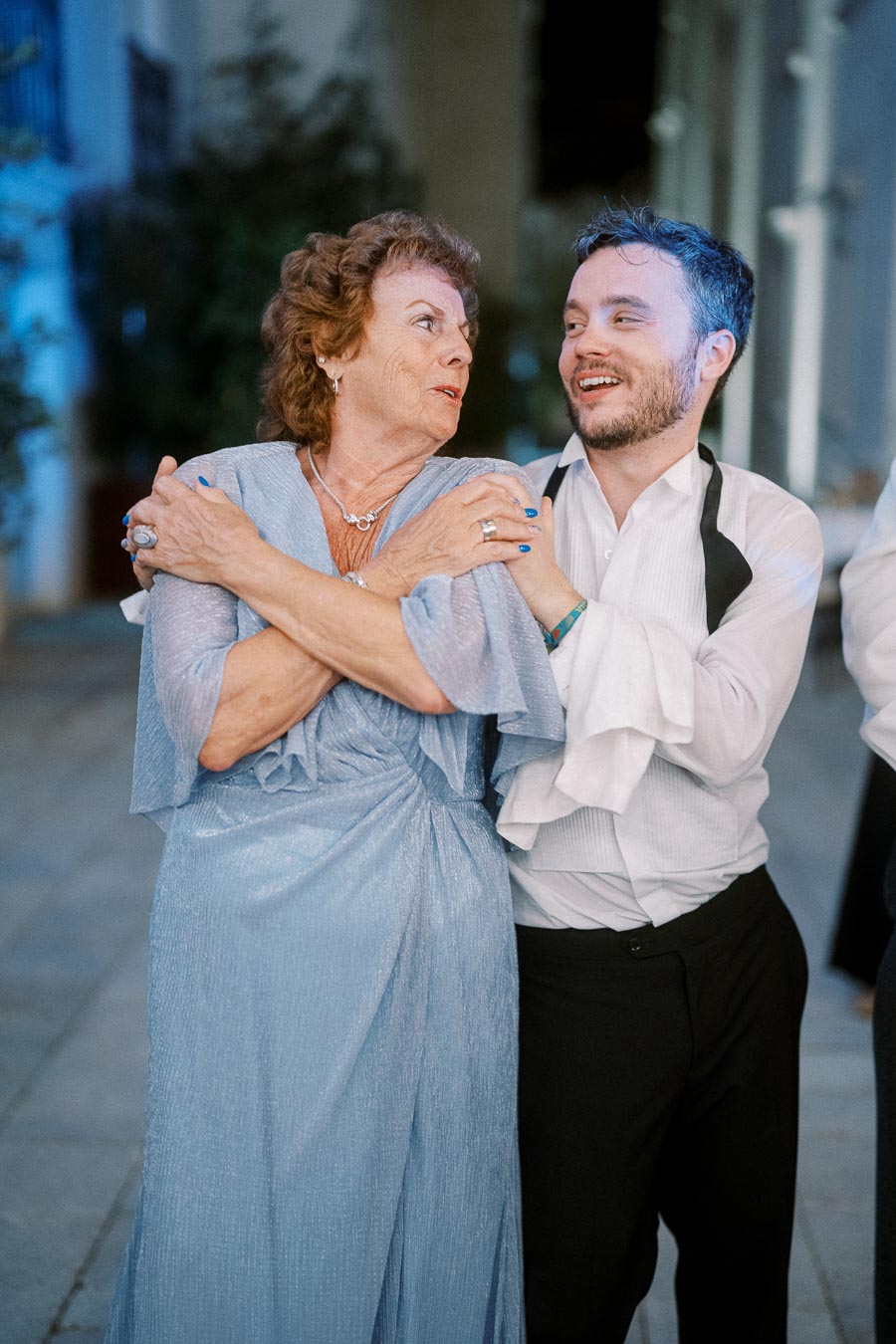 A joyful elderly woman in a shimmering blue dress dances with a smiling younger man in formal attire at an outdoor event, creating a heartwarming, intergenerational moment.