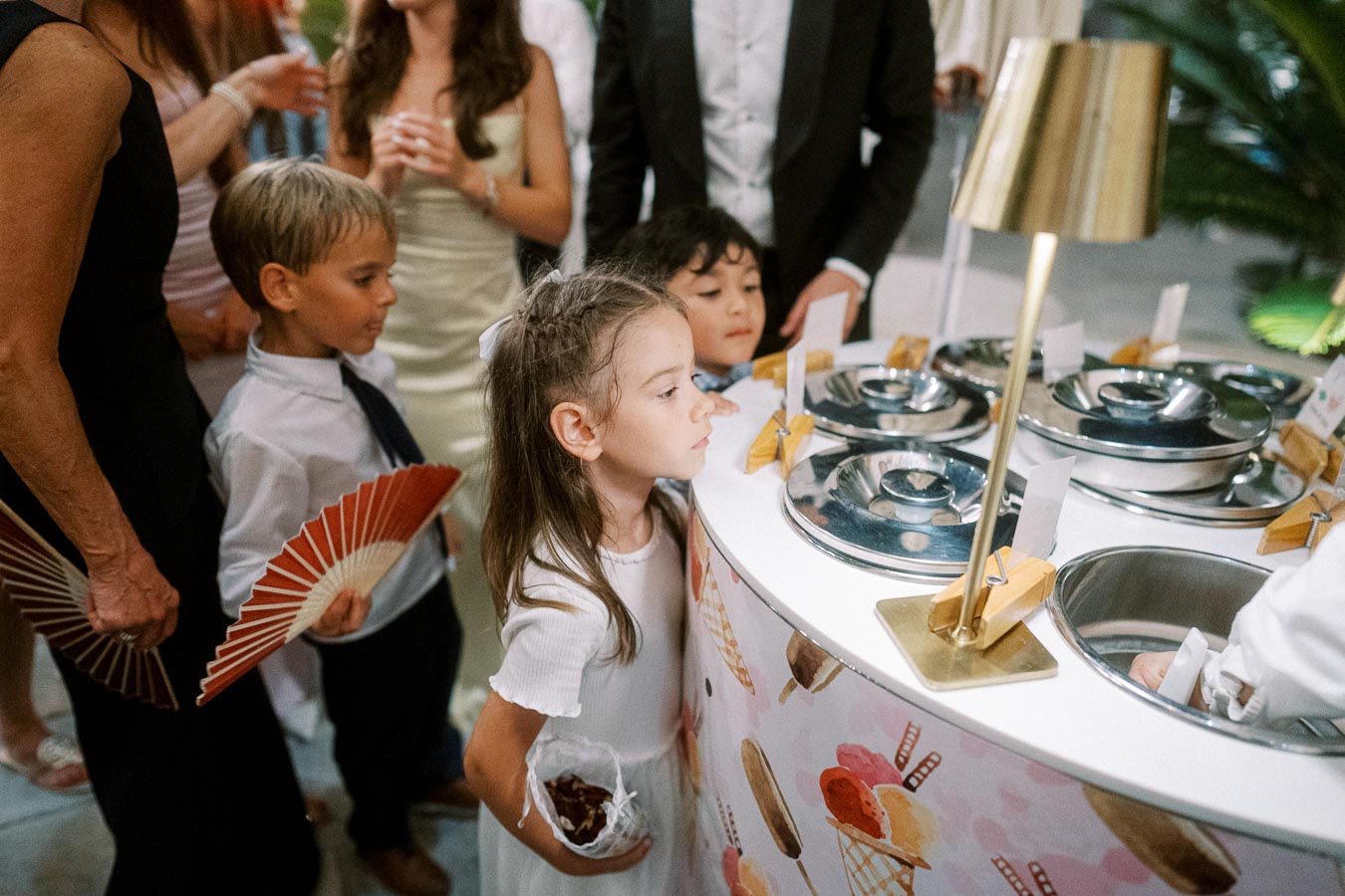 Children in formal attire eagerly looking at a gelato stand at an event, surrounded by adults holding fans. The scene captures a moment of excitement and anticipation.