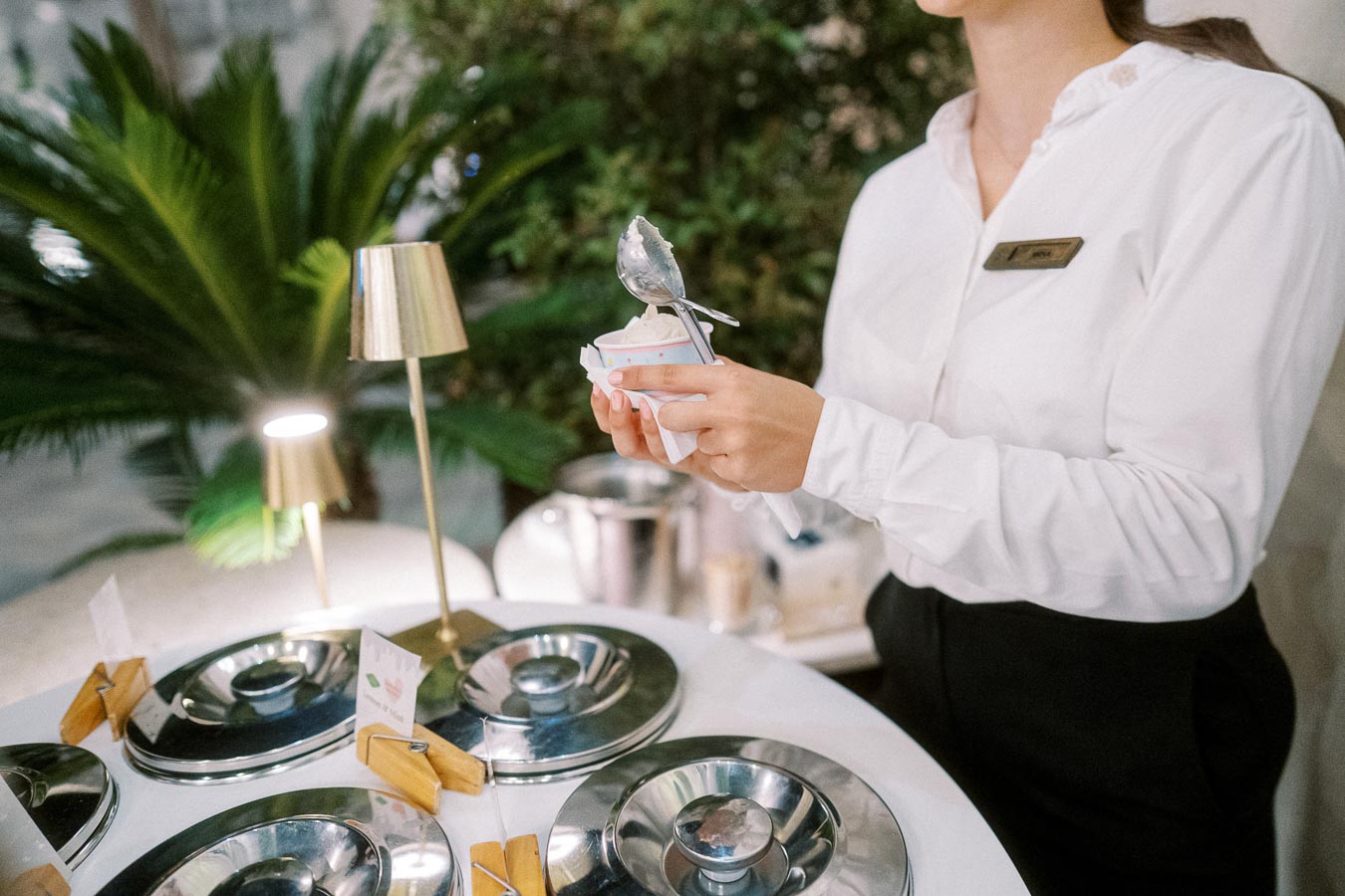 A hotel staff member in a white uniform is holding a cup of ice cream above a table with various metal lids and small signs, with lush greenery in the background.