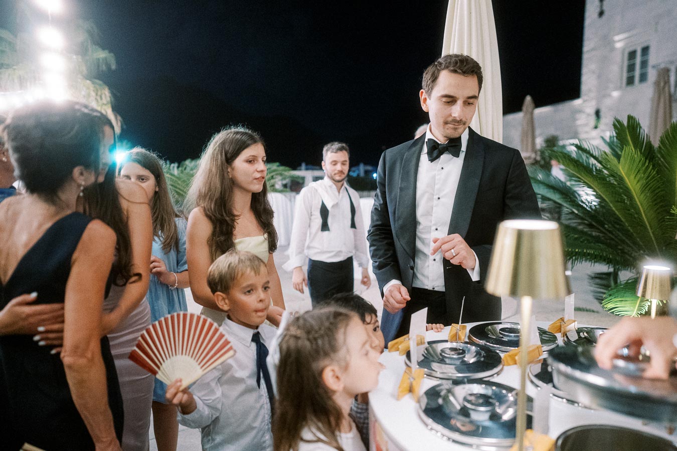 A group of elegantly dressed people gather around a dessert station at an outdoor evening event, with a man in a tuxedo serving ice cream to children.
