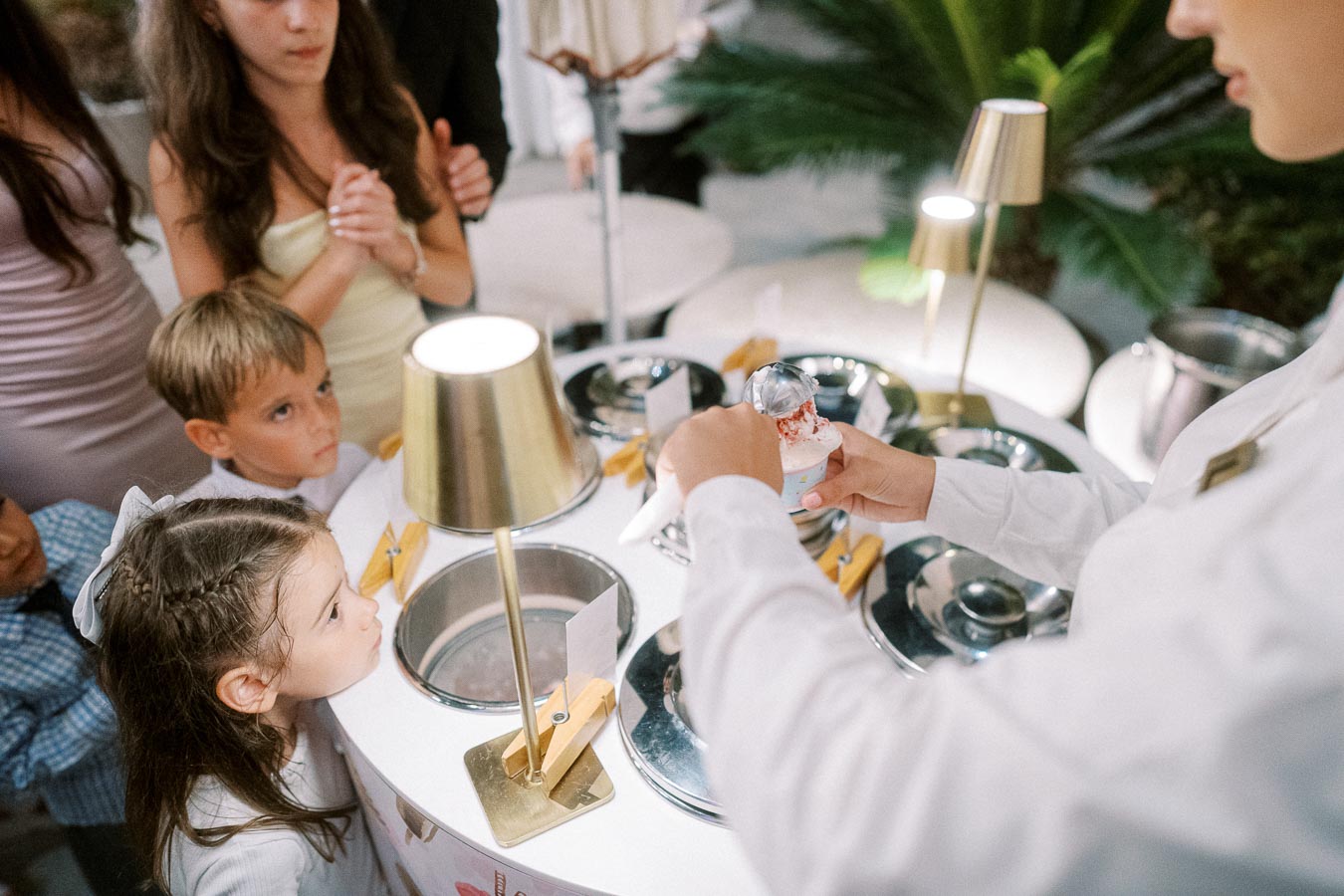 Children eagerly watching as a server prepares ice cream at an elegant dessert station, featuring multiple flavors and soft lighting, perfect for a family-friendly event.