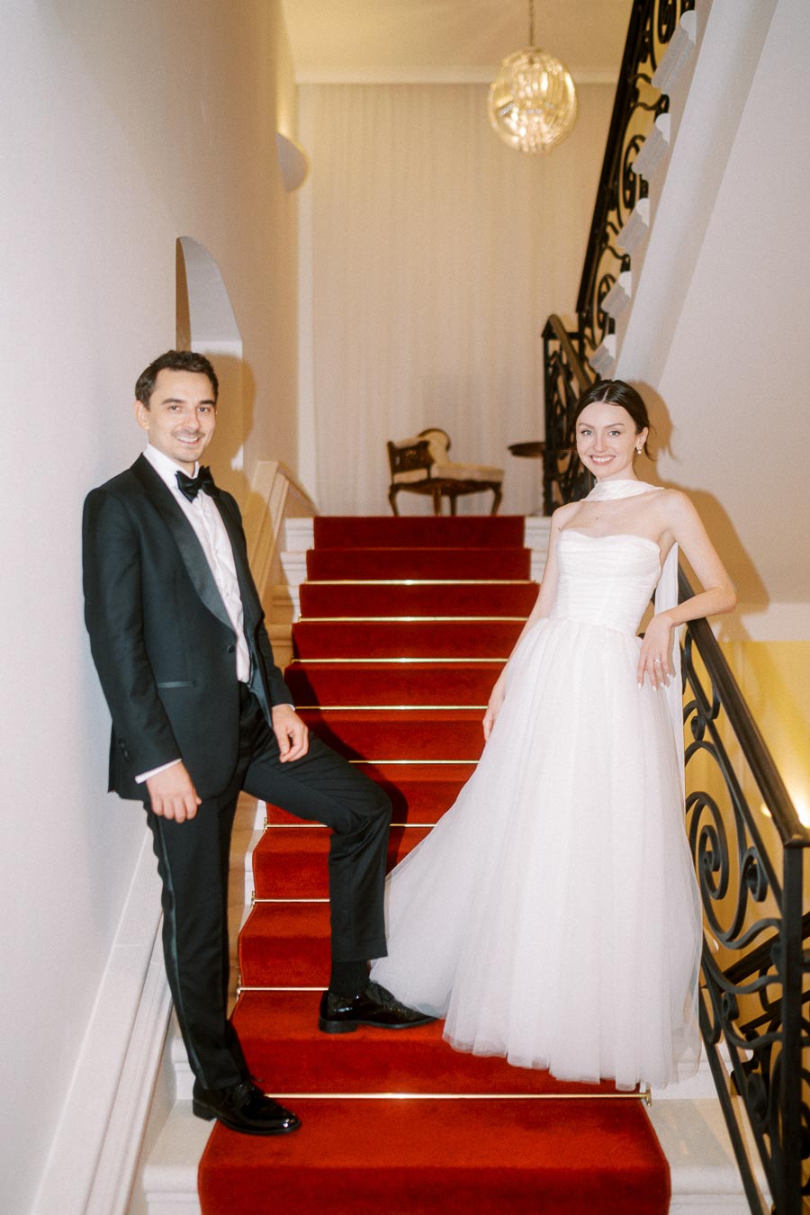 Elegant couple posing on a red carpeted staircase, with the man in a black tuxedo and the woman in a strapless white gown, under a hanging chandelier in a refined setting.