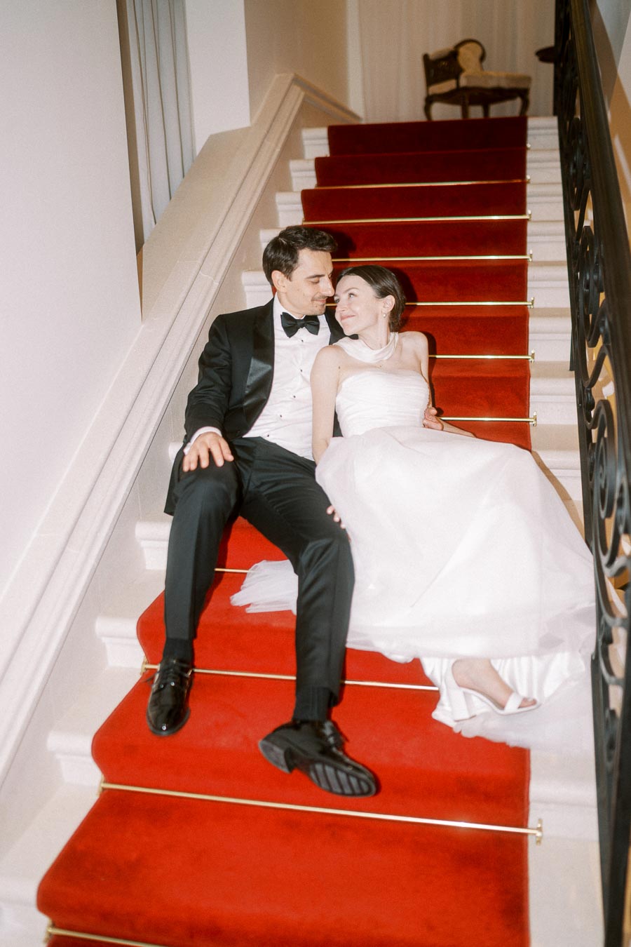 Bride and groom sitting on elegant red-carpeted staircase, sharing a romantic moment on their wedding day.
