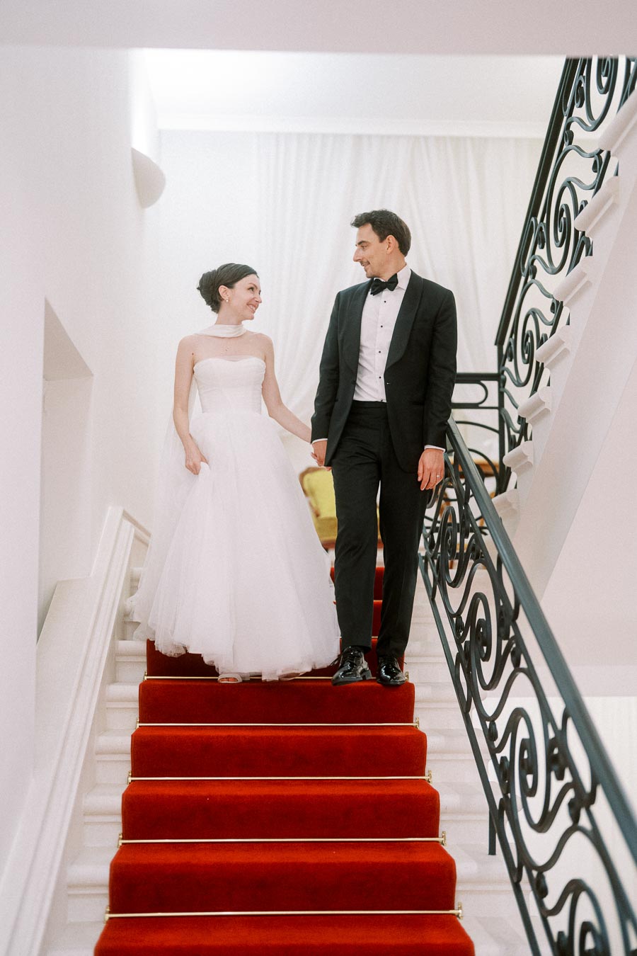 A bride in a white wedding dress and a groom in a black tuxedo walking down a staircase adorned with a red carpet, holding hands and smiling at each other in a beautifully decorated venue.