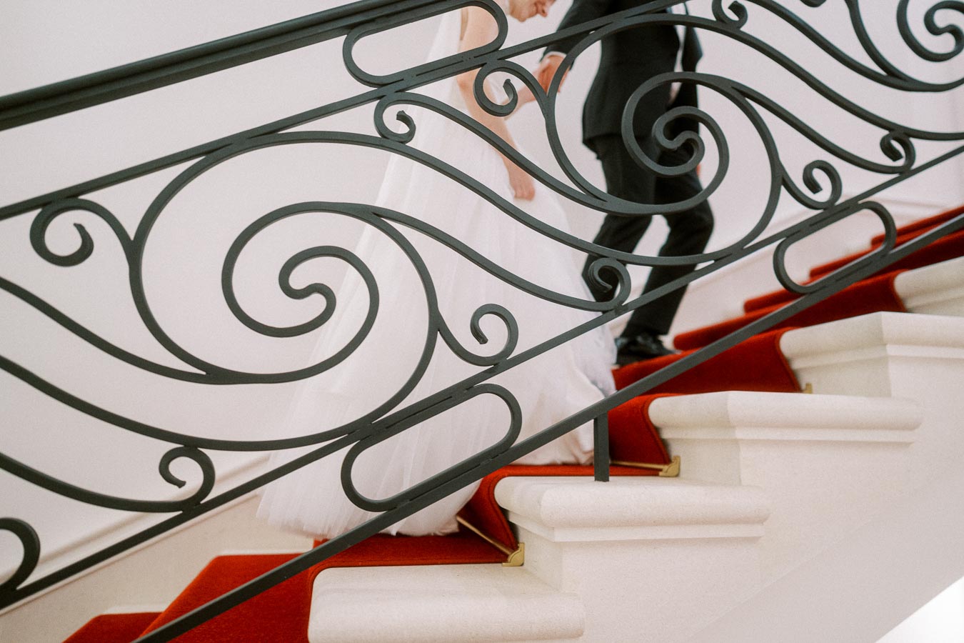 Wedding couple elegantly descending a grand staircase with ornate iron railing and red carpet in a luxurious setting.