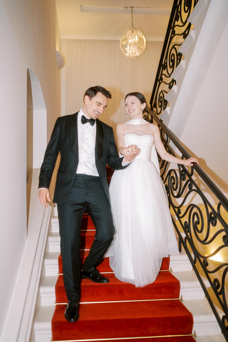 Elegant couple in formal attire walking down a decorated staircase, woman in a white gown, and man in a black tuxedo, holding hands and smiling, with a warm, luxurious setting in the background.