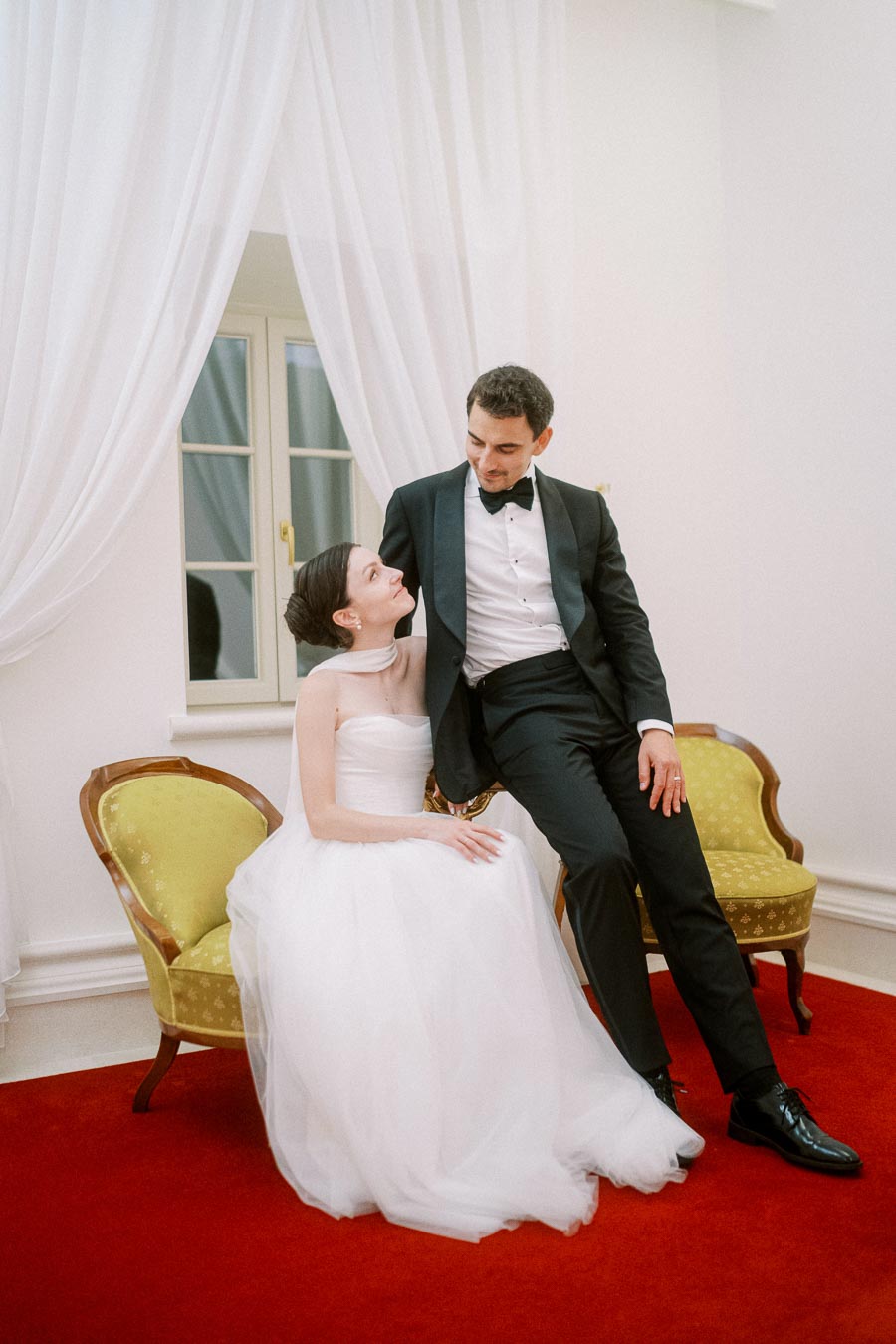 A bride in a white wedding gown sitting on a vintage chair looks up at a groom in a black tuxedo leaning on another matching chair, with elegant white drapes in the background.